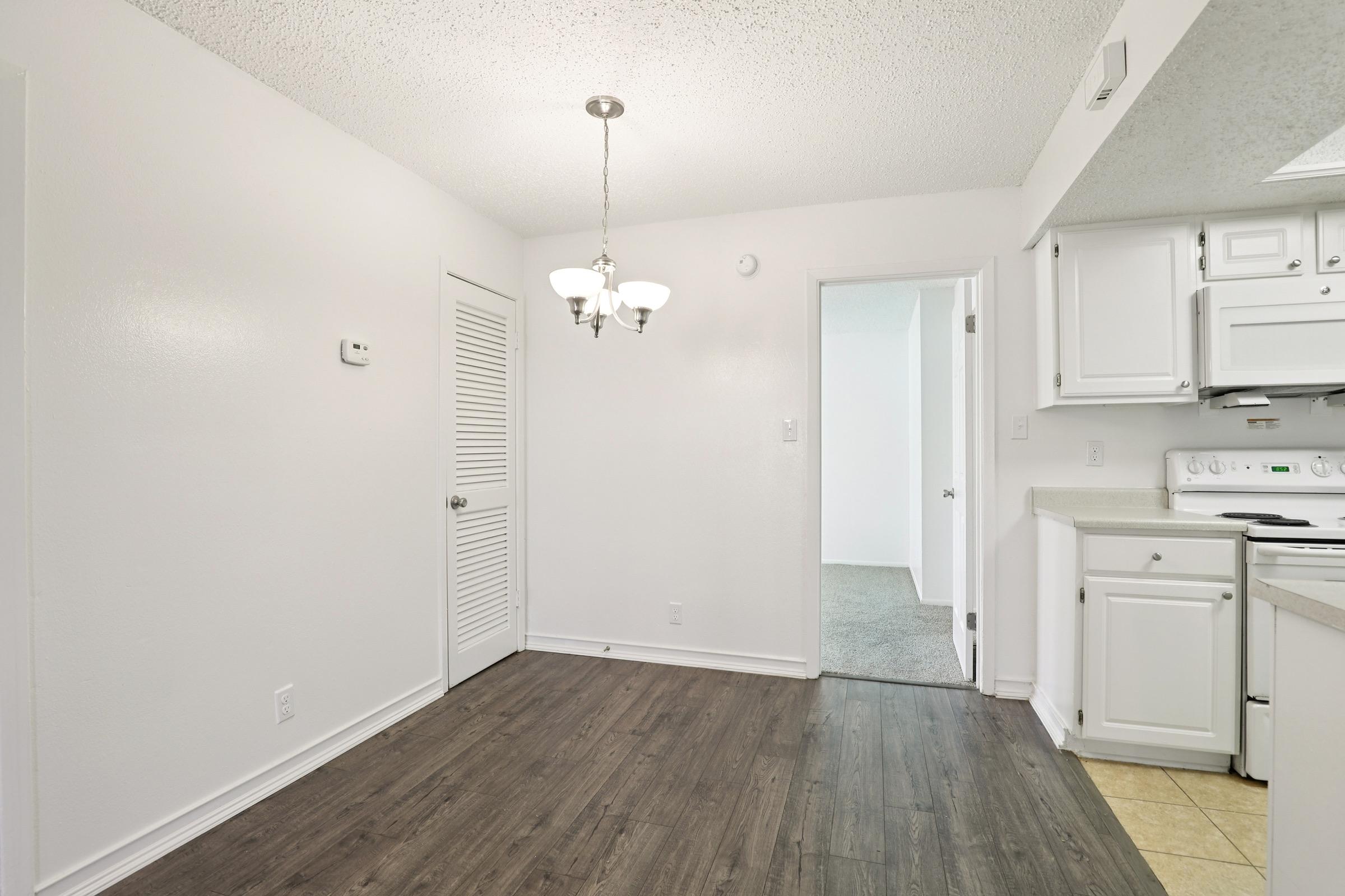 A bright, modern kitchen and dining area featuring white walls, a chandelier, and a combination of hardwood and tile flooring. A closed door leads to another room, while a doorway opens to the next space. Natural light fills the area, creating an inviting atmosphere.