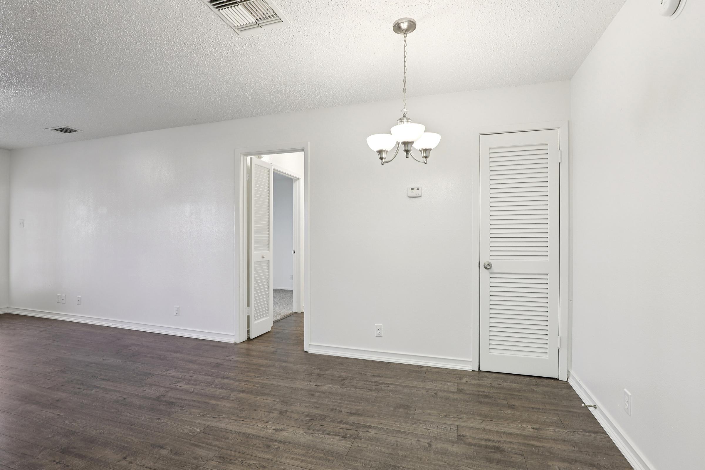 Interior view of a room featuring light-colored walls and a textured ceiling. A ceiling light fixture hangs centrally, and there is a closed door on the right leading to a closet. An open doorway on the left leads to another room. The floor is covered with a wooden finish, creating a warm and inviting atmosphere.