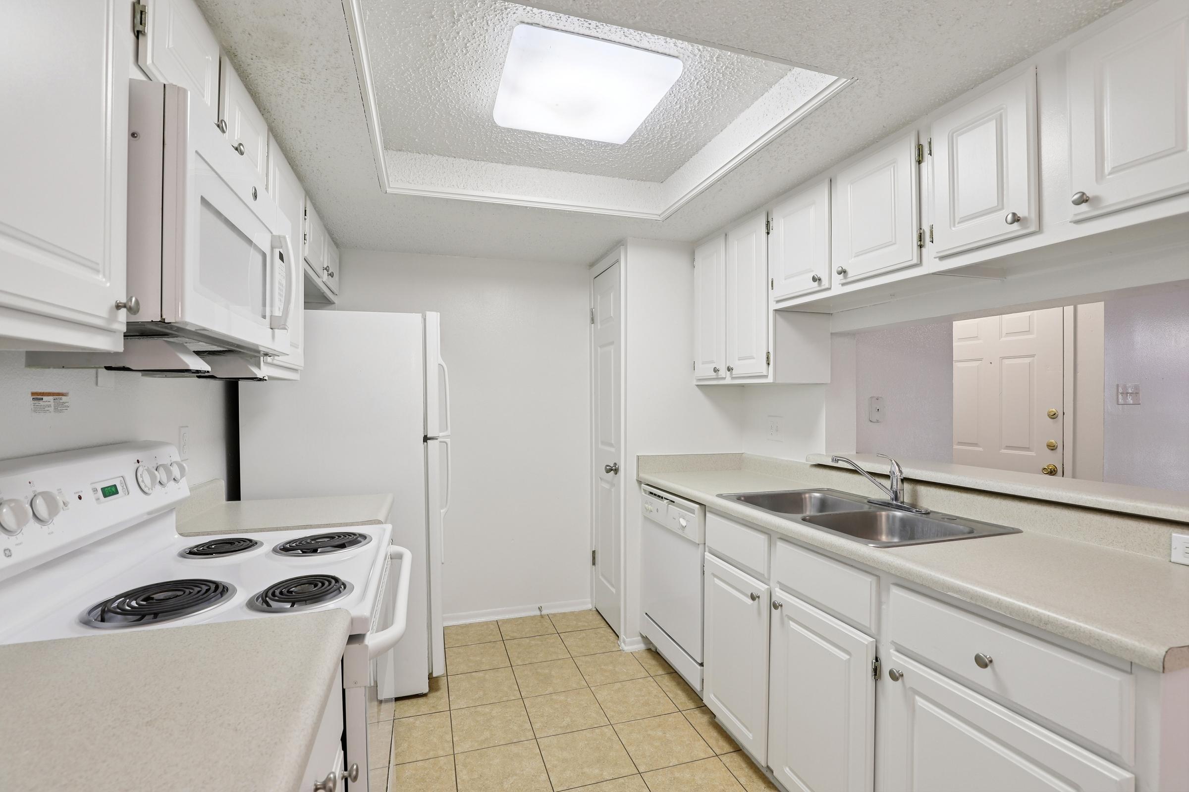 A clean and modern kitchen featuring white cabinetry, a large sink, and white appliances including a stove, microwave, and refrigerator. The floor is tiled in light colors, and the space is illuminated by a ceiling light fixture. The walls are painted in neutral tones, creating a bright and airy atmosphere.