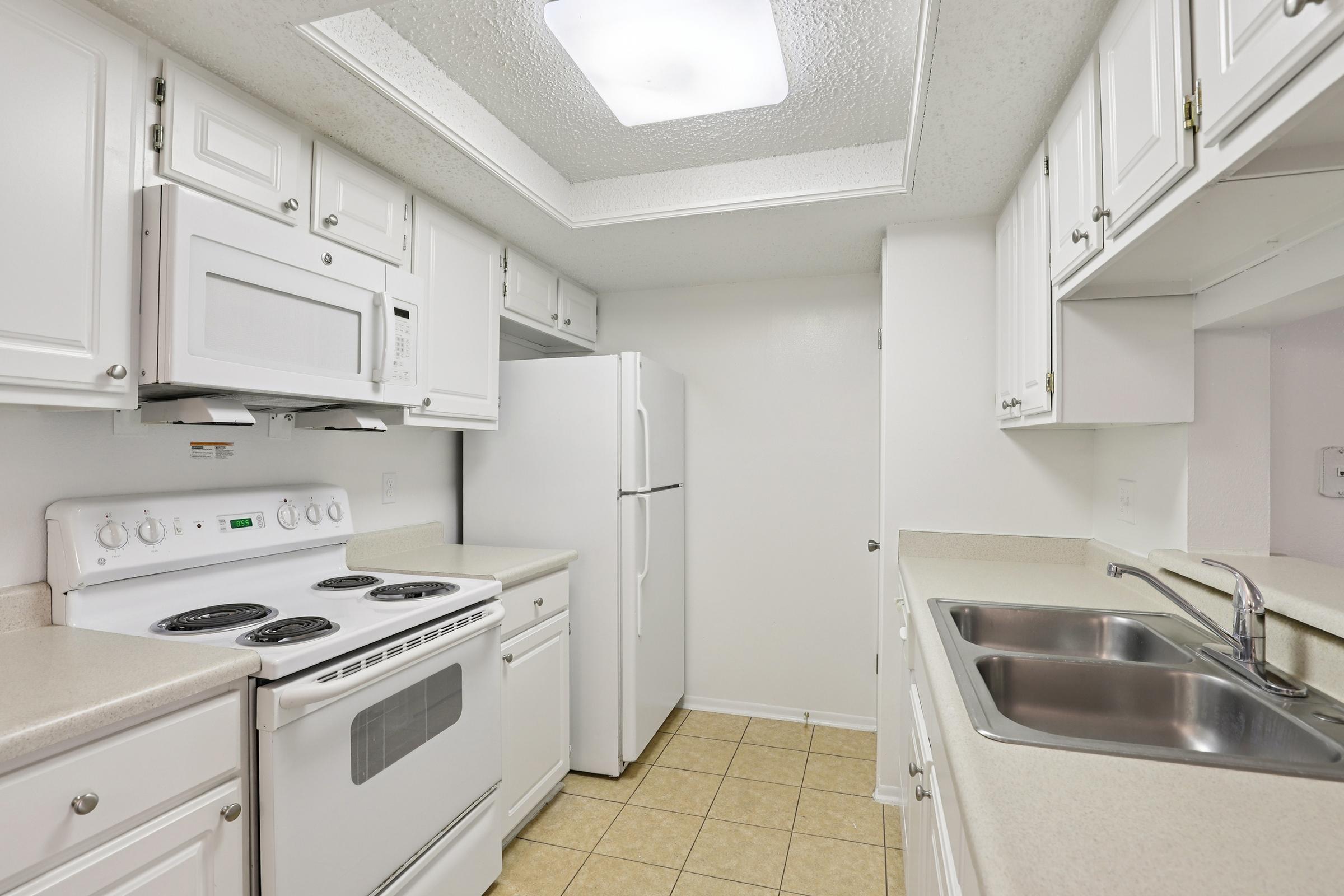A small, modern kitchen featuring white cabinetry, a white stove and microwave, and a double sink. The countertops are light-colored and the flooring is tiled. Bright overhead lighting illuminates the space, creating a clean and functional environment. A refrigerator is also visible along the wall.