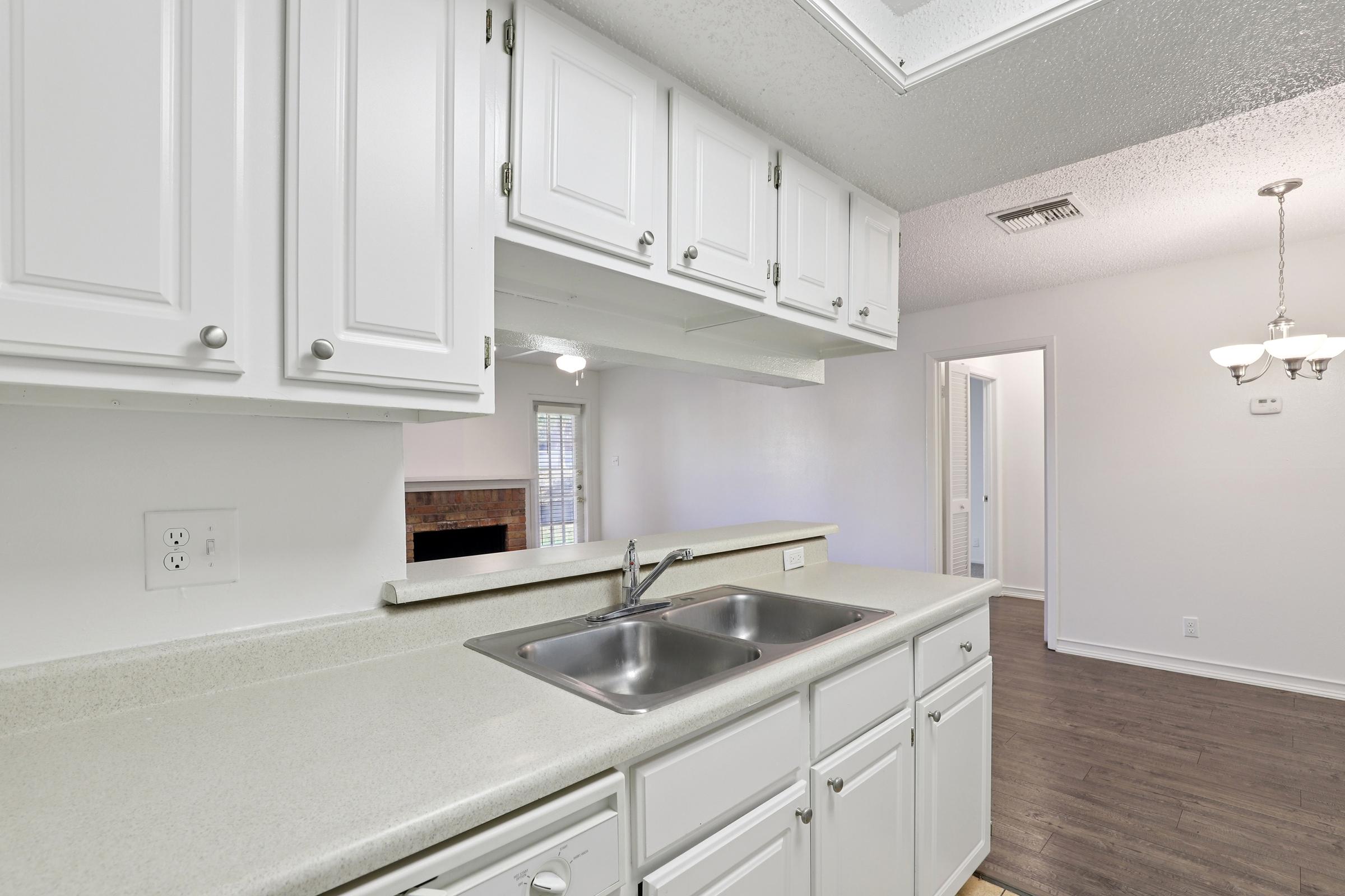 A modern kitchen featuring white cabinets, a double sink, and a countertop. Natural light floods the space, illuminating the walls and showcasing a view into a nearby room with a door. The flooring is dark wood, and there is a light fixture hanging above the dining area.