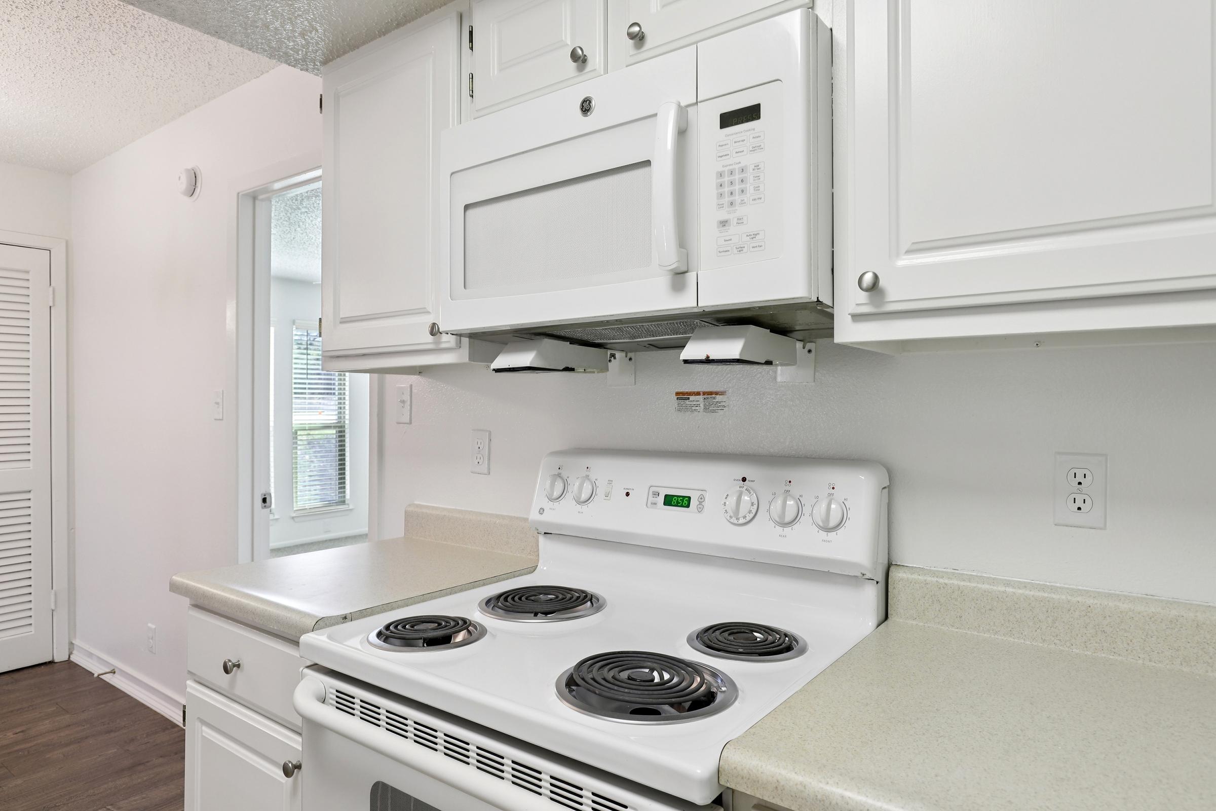 A clean, modern kitchen featuring white cabinets, a microwave above a white stove with four burners, and a light-colored countertop. The room has a window visible, allowing natural light to enter the space. The overall design is bright and inviting.