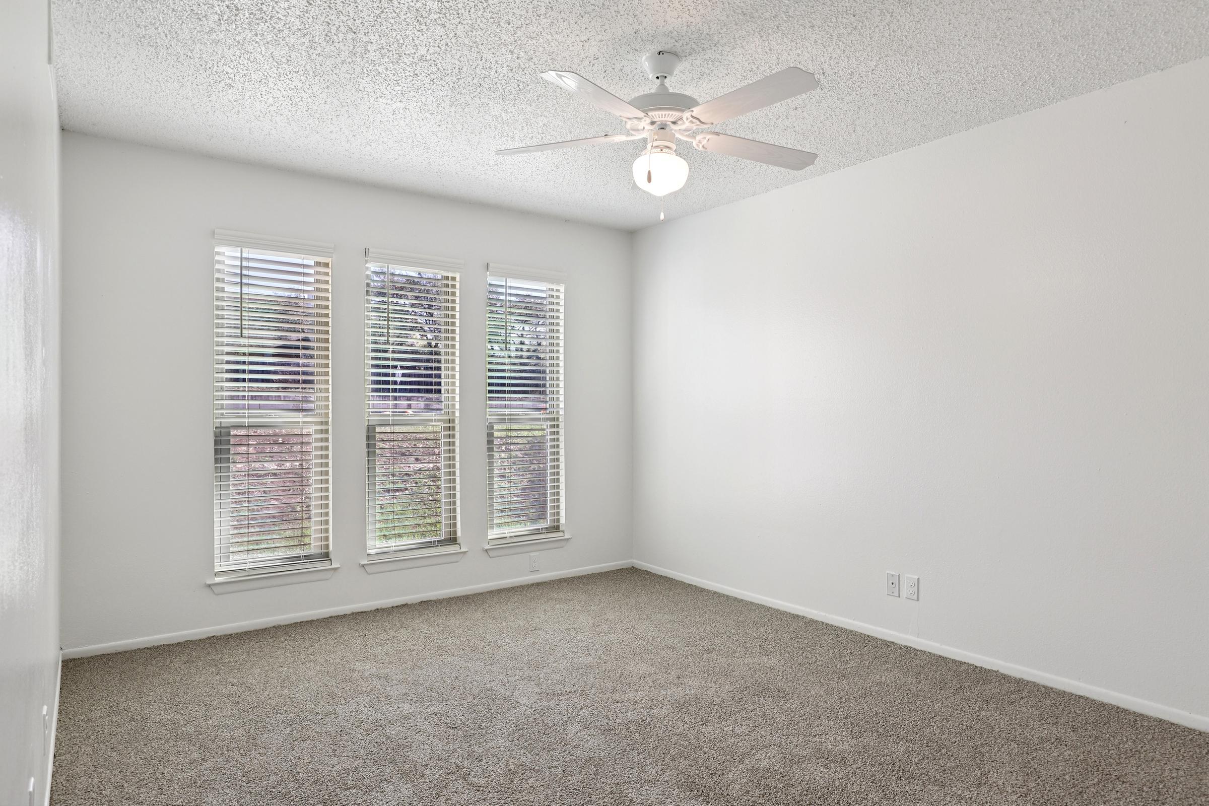 A bright, empty room with light-colored walls and a ceiling fan. There are two windows with blinds allowing natural light to enter, and the floor is covered in beige carpet. The room appears spacious and uncluttered, providing a clean and versatile living space.
