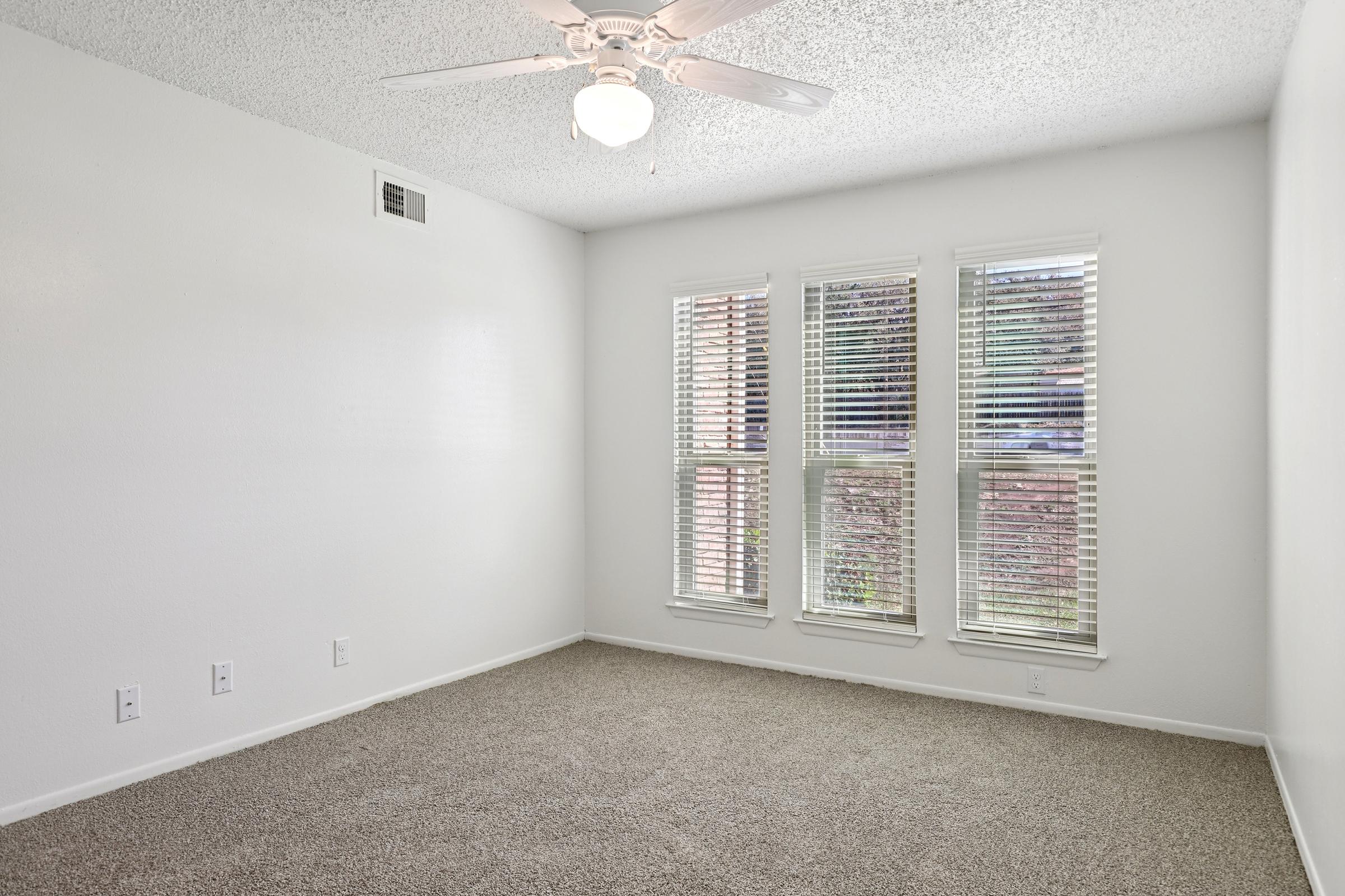 A well-lit, empty room featuring beige carpet, a ceiling fan, and three large windows with blinds. The walls are painted white, and there are electrical outlets visible. The overall atmosphere is bright and clean, suggesting a fresh and minimalist space.