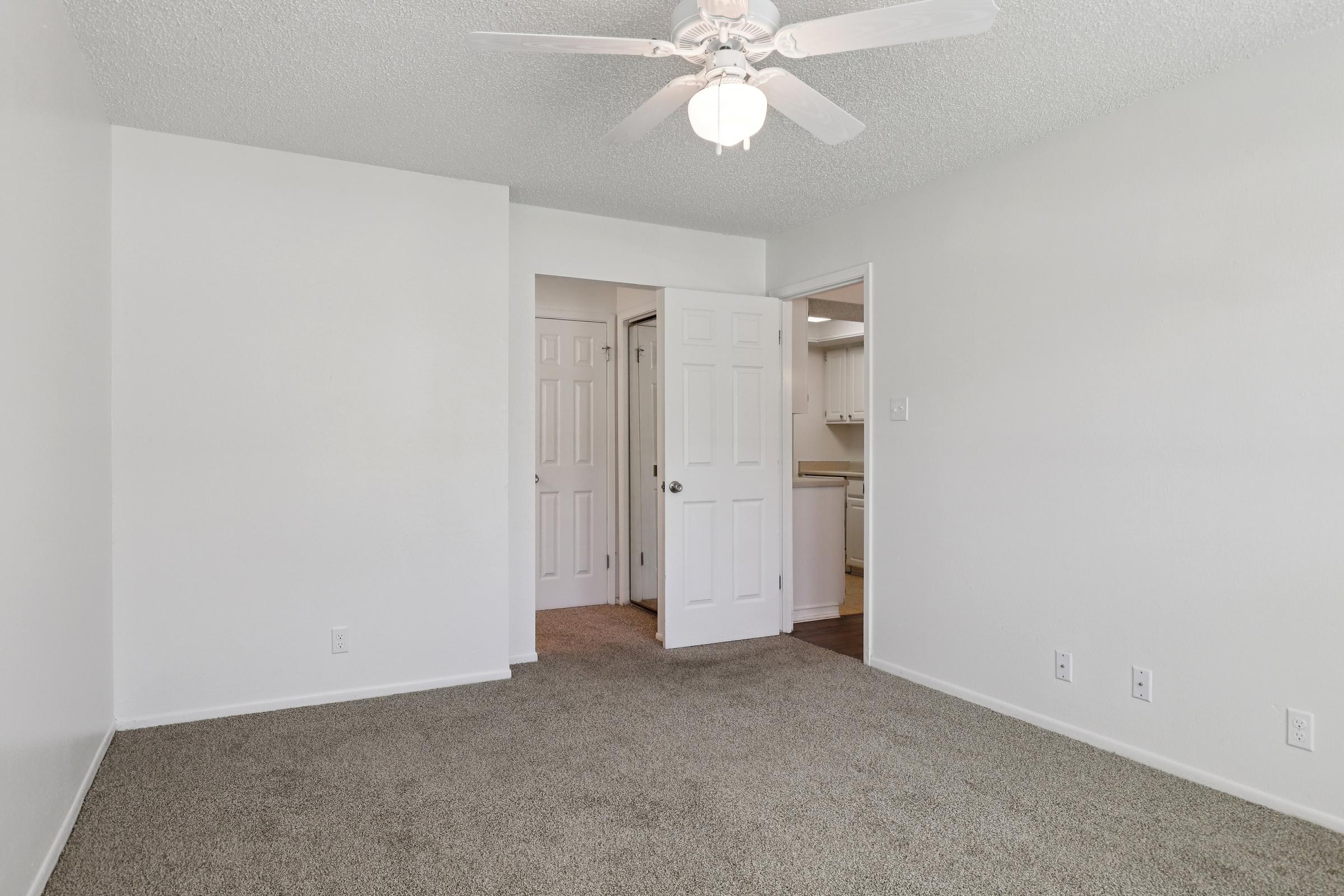 A well-lit, vacant room featuring beige carpet and white walls. A ceiling fan is visible, and there are two doors leading to other areas. One door appears to lead to a kitchen. The room is neat and simple, creating an open and spacious atmosphere.