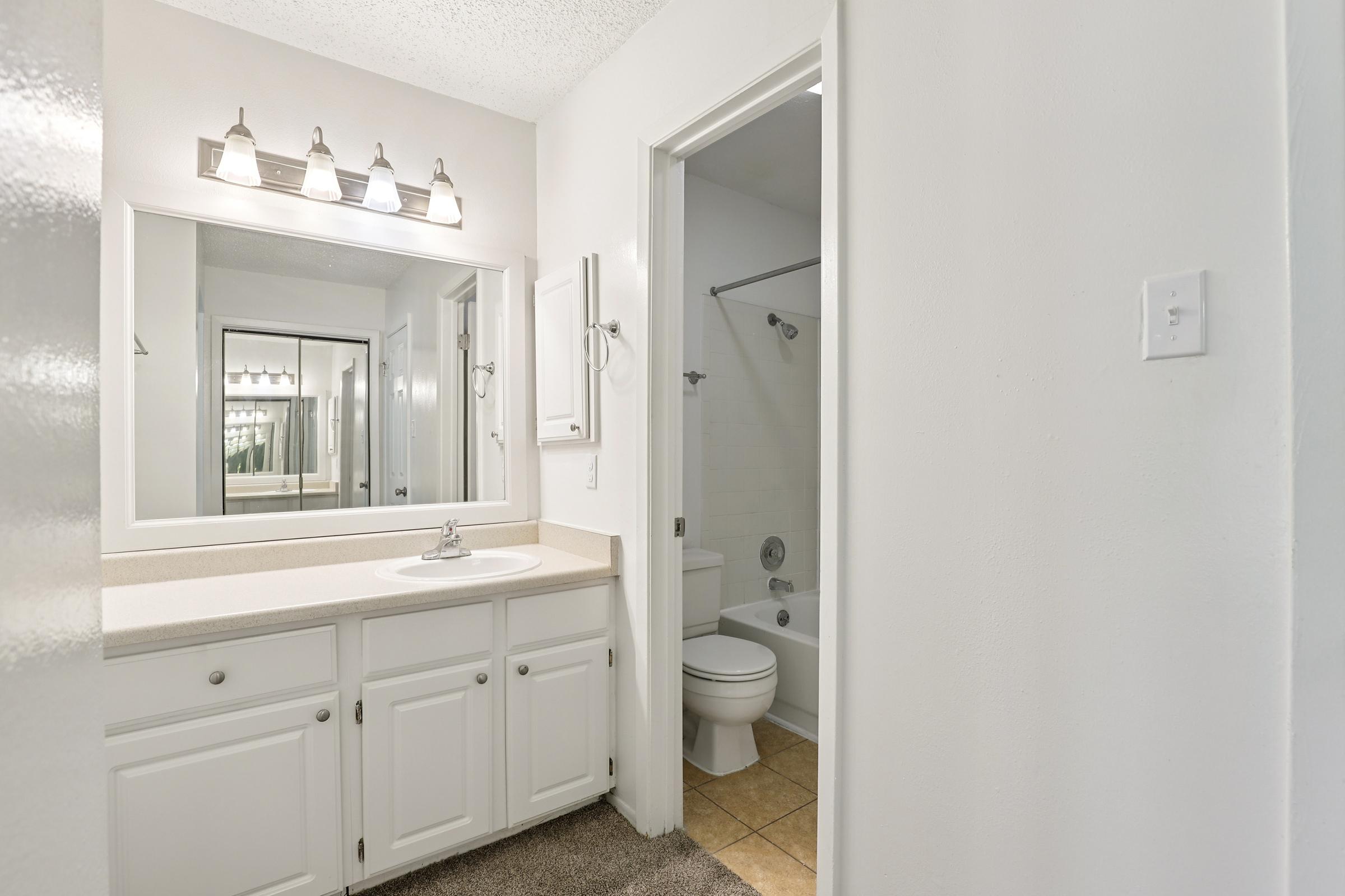 A clean, well-lit bathroom featuring a white vanity with a sink, large mirror, and wall-mounted light fixtures. There is a shower-tub combination with a curtain, and beige tile flooring. A door leads to a small room, and the walls are painted white, giving a spacious and airy feel to the space.