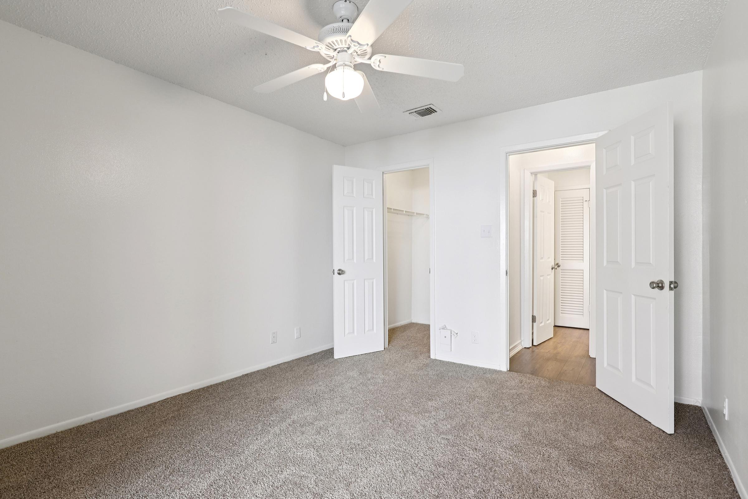 A bright, empty room with light-colored walls and carpeted flooring. It features a ceiling fan, two open white doors leading to closets and an adjacent space, and a neutral color scheme, creating a clean and spacious atmosphere.