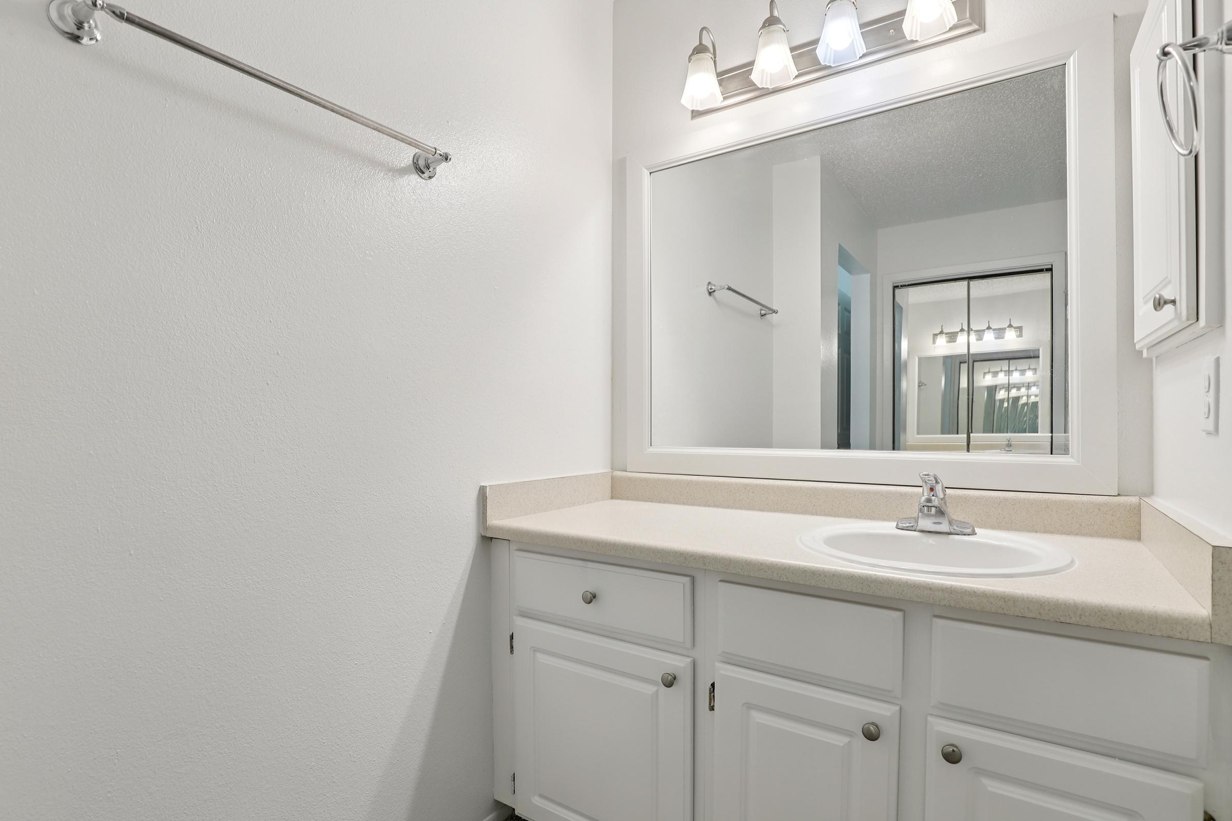 A clean, minimalistic bathroom featuring a white vanity with a sink, a large mirror above, and light fixtures. The walls are painted white and there is a towel rack to the left. The space appears bright and modern, with a simple countertop and cabinetry below the sink.