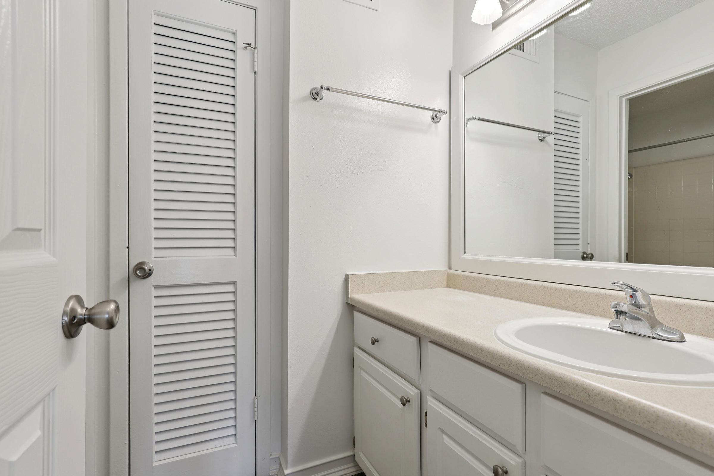 A clean and modern bathroom featuring a large mirror above a light-colored countertop with a sink. There is a cabinet underneath the sink and a door leading to a closet or bathroom area on the left. The walls are painted white, and there is a towel rack above the countertop.