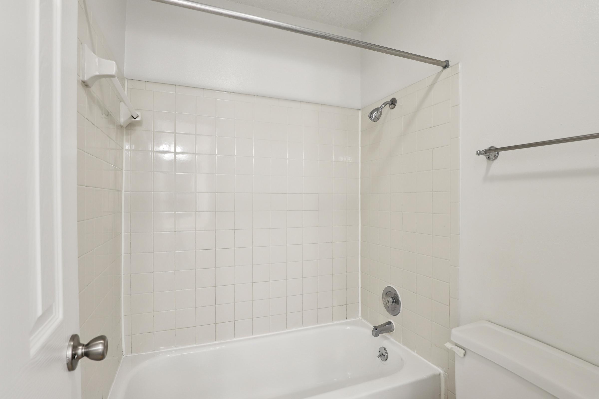 A clean, empty bathroom featuring a white tiled shower area, a bathtub, a shower curtain rod, a wall-mounted towel rack, and a toilet. The walls are painted white, creating a bright and spacious appearance. The overall design is simple and modern.