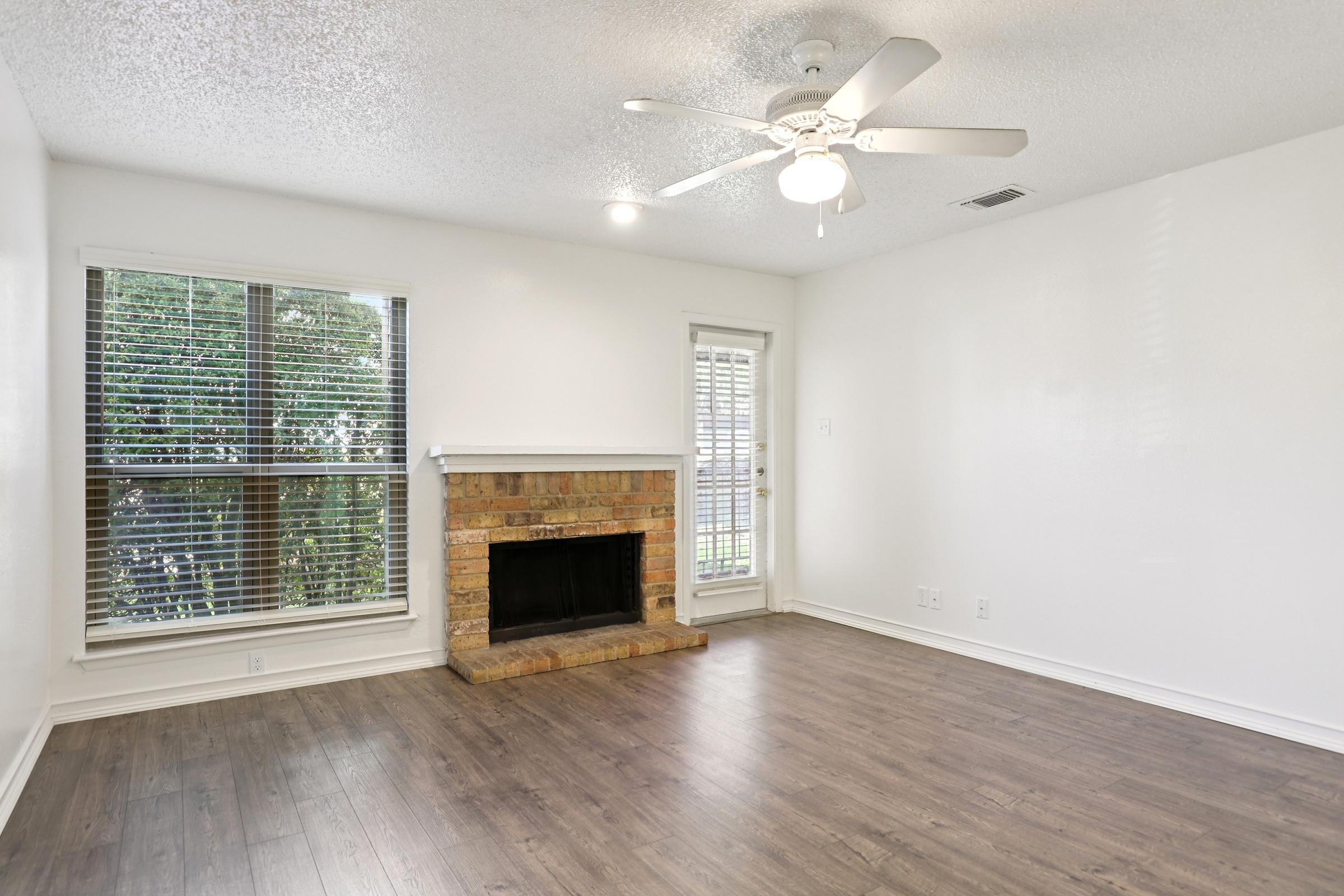 Spacious empty living room featuring a brick fireplace, ceiling fan, and large windows with blinds. Light-colored walls and hardwood flooring create a bright and inviting atmosphere. A door leads to an outdoor area, enhancing natural light in the room.