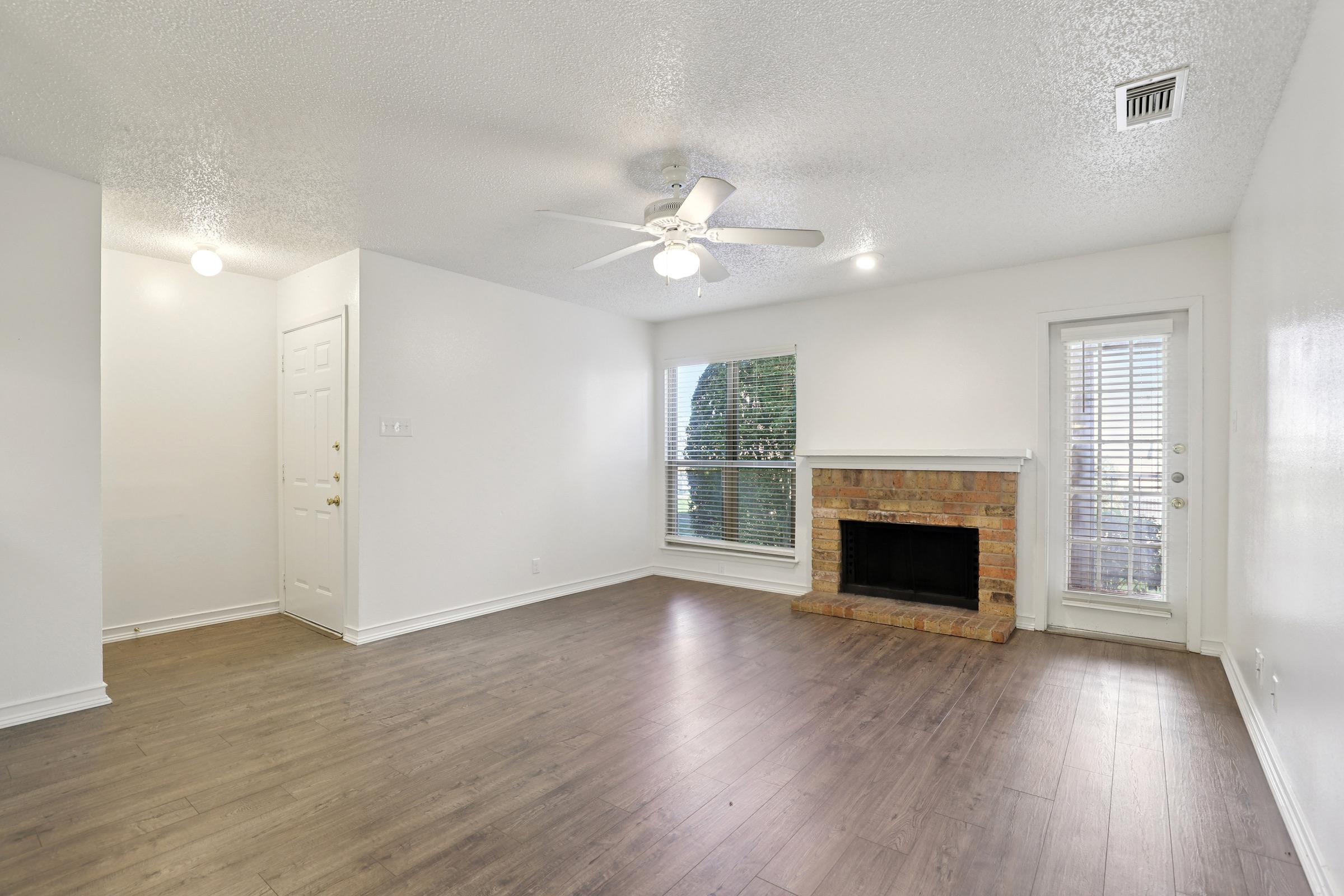 Empty living room with a ceiling fan, large window, and brick fireplace. Light-colored walls and wood-like flooring create a spacious feel. A door leads outside, and bright lights illuminate the room, making it inviting and well-lit.