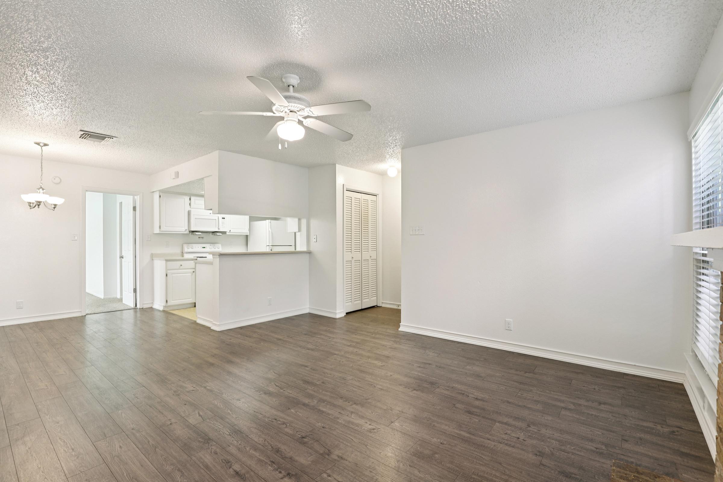 A well-lit, modern living room featuring a ceiling fan, light-colored walls, and dark wood flooring. In the background, there's a kitchen area with white cabinets and appliances. A doorway leads to another room, and there are closed closet doors on one wall. The space is bright and inviting.