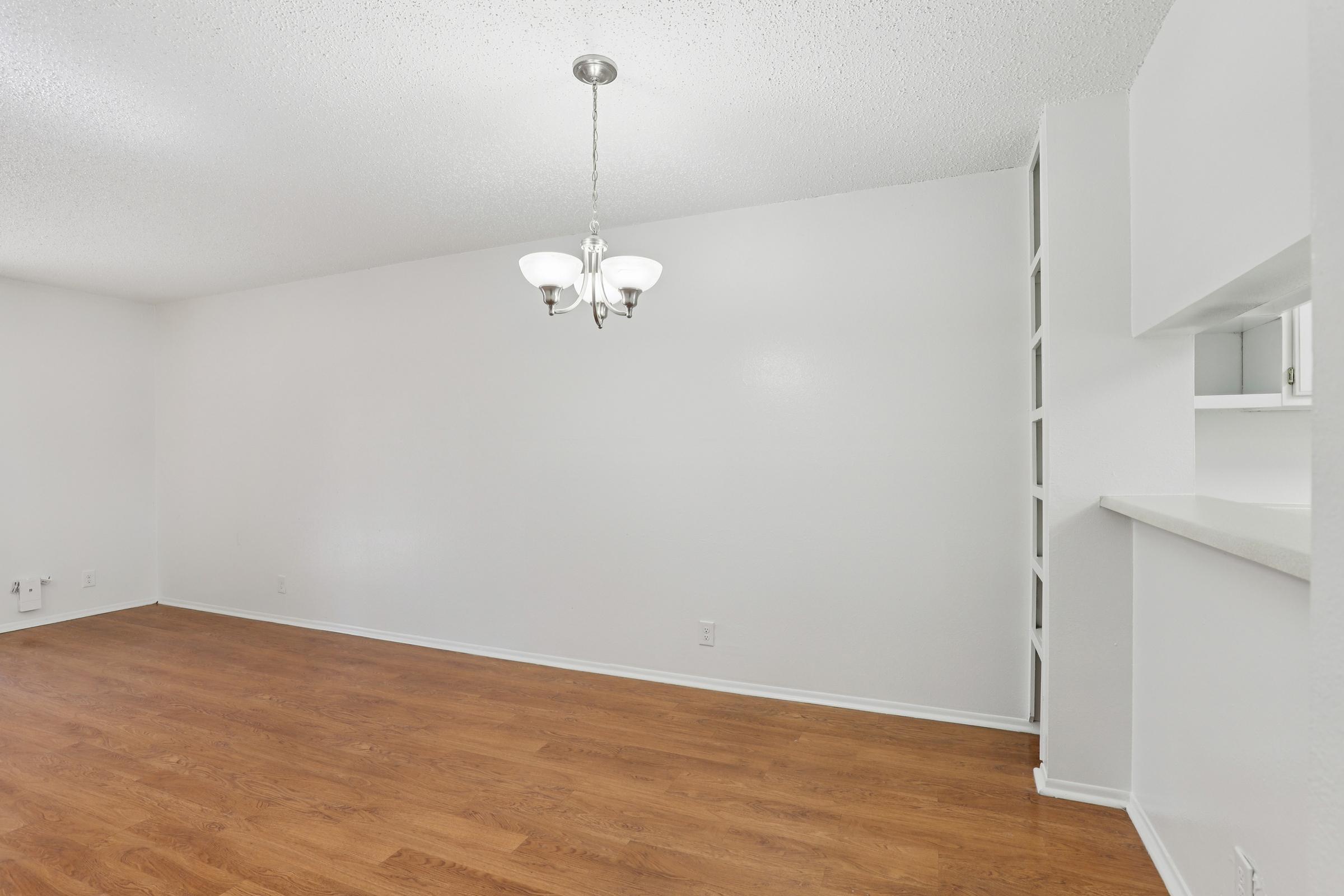 Spacious interior of a living room featuring light-colored walls, hardwood flooring, and a ceiling chandelier. There is a small open shelving unit built into the wall on the right side, and the room is empty, highlighting its potential for decoration and furniture arrangement.