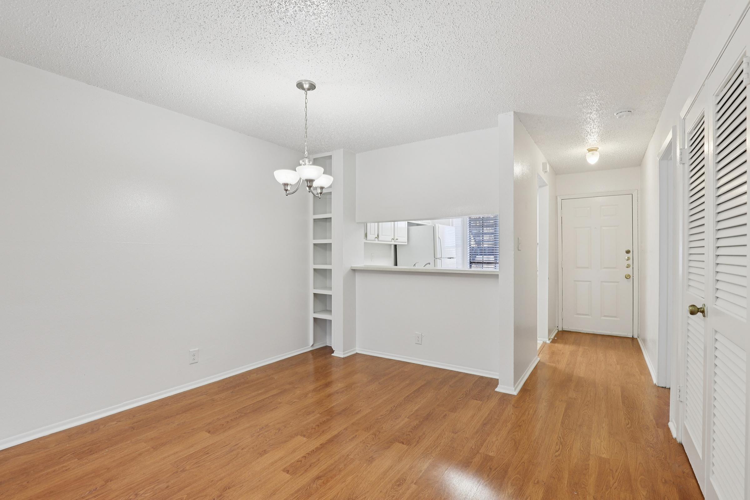 A bright, open living space featuring light wood flooring, a white textured ceiling, and walls. There's a small chandelier hanging above, and a doorway leads to the entrance. To the left, a shelf is built into the wall, and a window can be seen through the entryway, allowing natural light into the room.