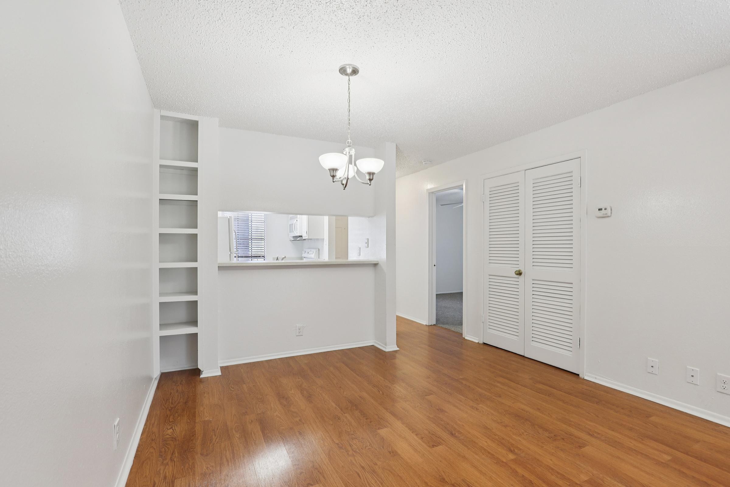 Empty living room with light wooden flooring, white walls, and a ceiling light fixture. There is a built-in shelf on one side and a doorway leading to another room. A small bar area is visible in the background, connecting to a kitchen. The space is well-lit and has a clean, modern look.