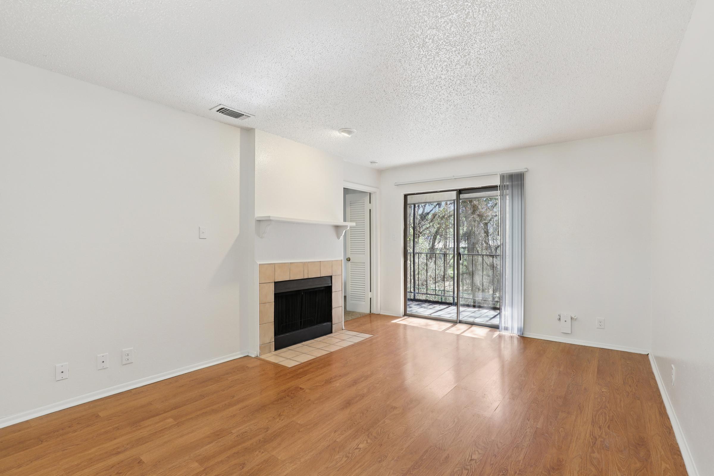 A spacious living room featuring a light-colored wall, hardwood flooring, a tiled fireplace, and a large window with a sliding door that leads to a balcony. Natural light floods the room, creating a bright and airy atmosphere. Minimalist design with no furniture visible.