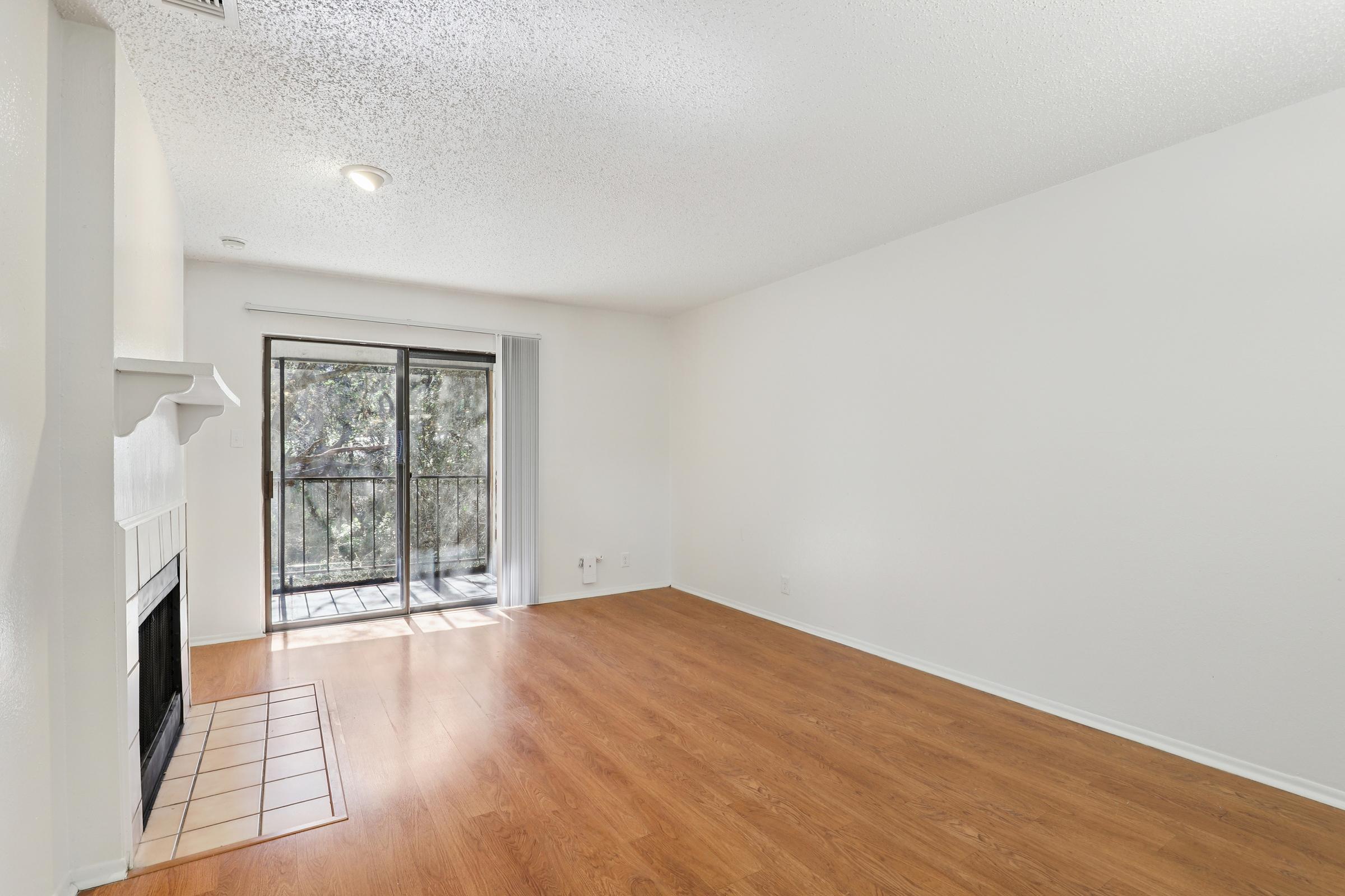 A spacious, empty living room featuring a wooden floor, a fireplace on the left, and a sliding glass door leading to a balcony. Natural light streams in through the door, illuminating the white walls and ceiling. The room has a simple, clean aesthetic, ideal for personalization.
