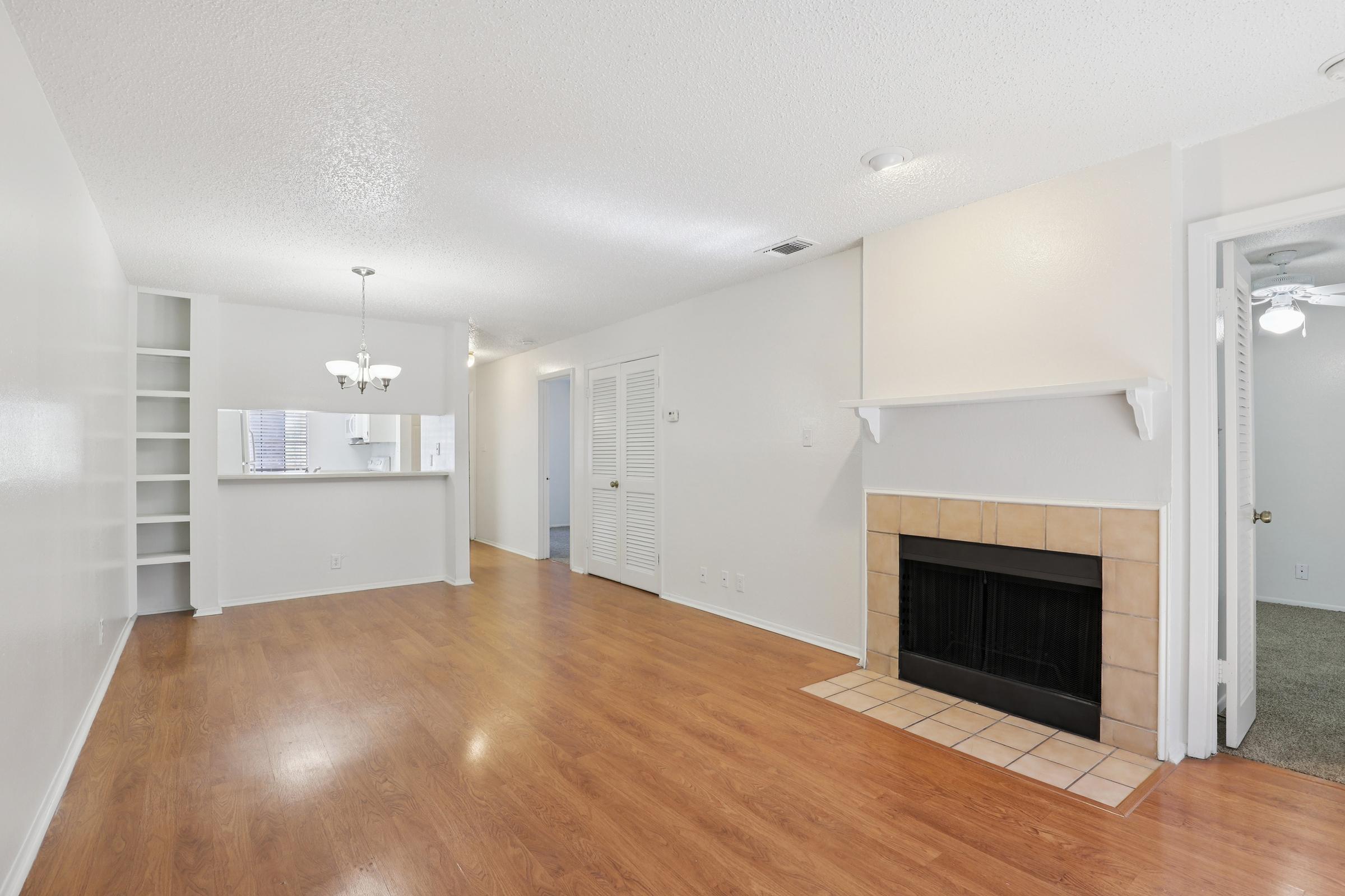 A spacious living room featuring wood-style flooring, a cozy fireplace, and a large window. The area includes built-in shelving and a dining area with a chandelier. Doors lead to a bedroom and a bathroom, while walls are painted in a light color, creating an open and inviting atmosphere.