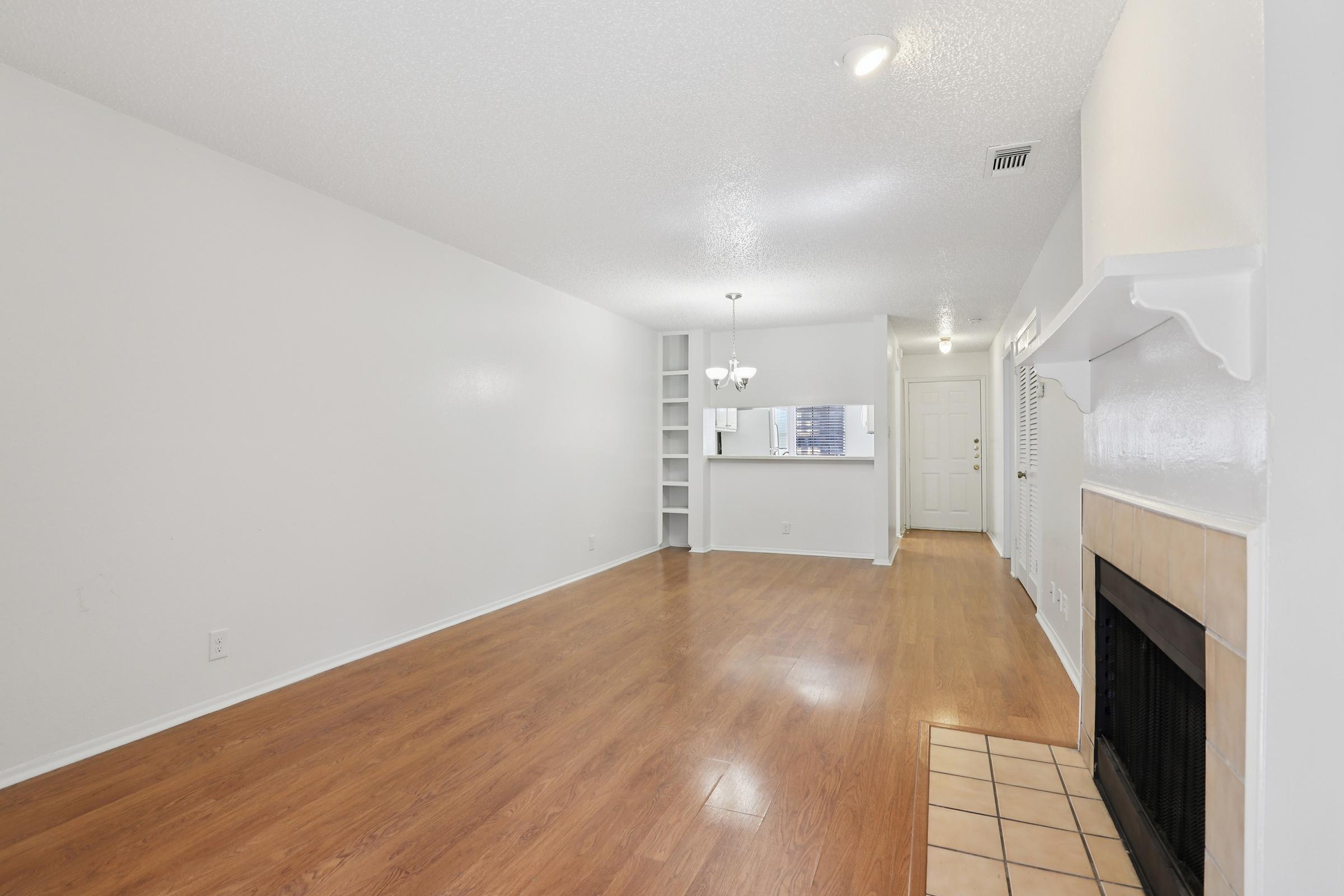 A spacious, well-lit living area with light hardwood floors and white walls. There's a small fireplace on one side and a chandelier hanging from the ceiling. In the background, a doorway leads to another room, and a built-in shelf is visible against the wall. The overall atmosphere is bright and inviting.