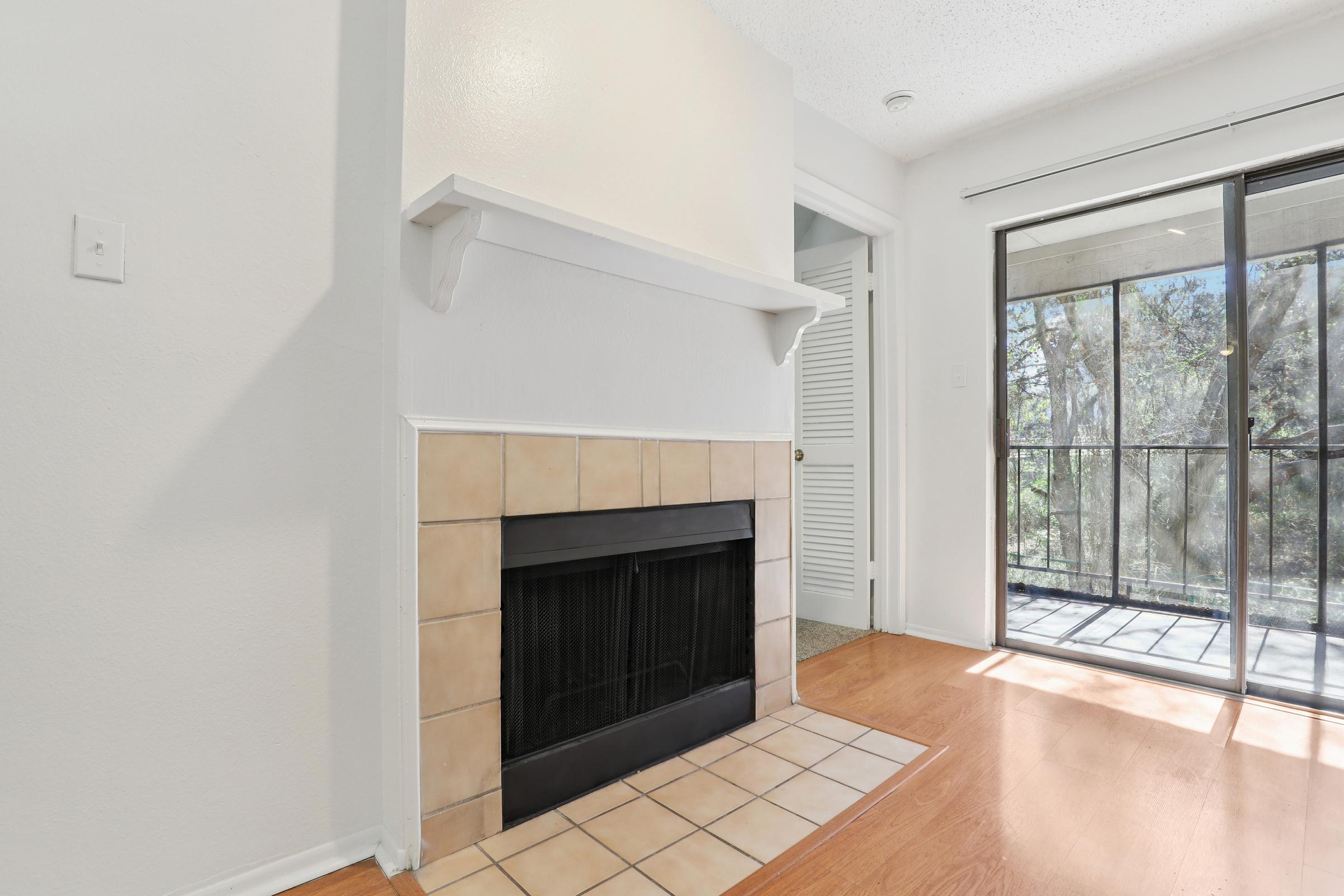 A cozy interior featuring a tiled fireplace with a white mantel, complemented by light-colored walls. To the right, a sliding glass door leads to a balcony or outdoor space, framed by light wood flooring. The space feels bright and inviting, creating a warm atmosphere.