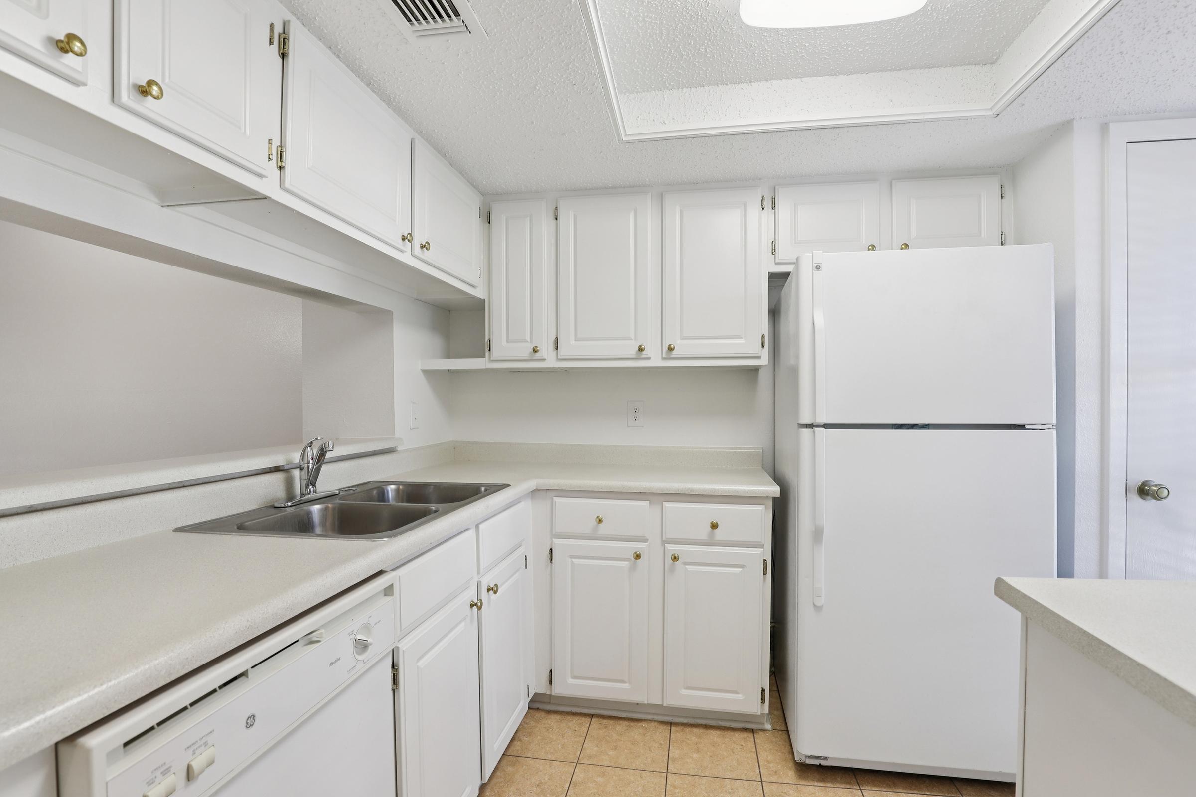 A clean and modern kitchen featuring white cabinetry, a double sink, a dishwasher, and a large white refrigerator. The tile floor adds to the bright and airy feel, with ample counter space and shelving for storage. The layout is functional and well-organized.