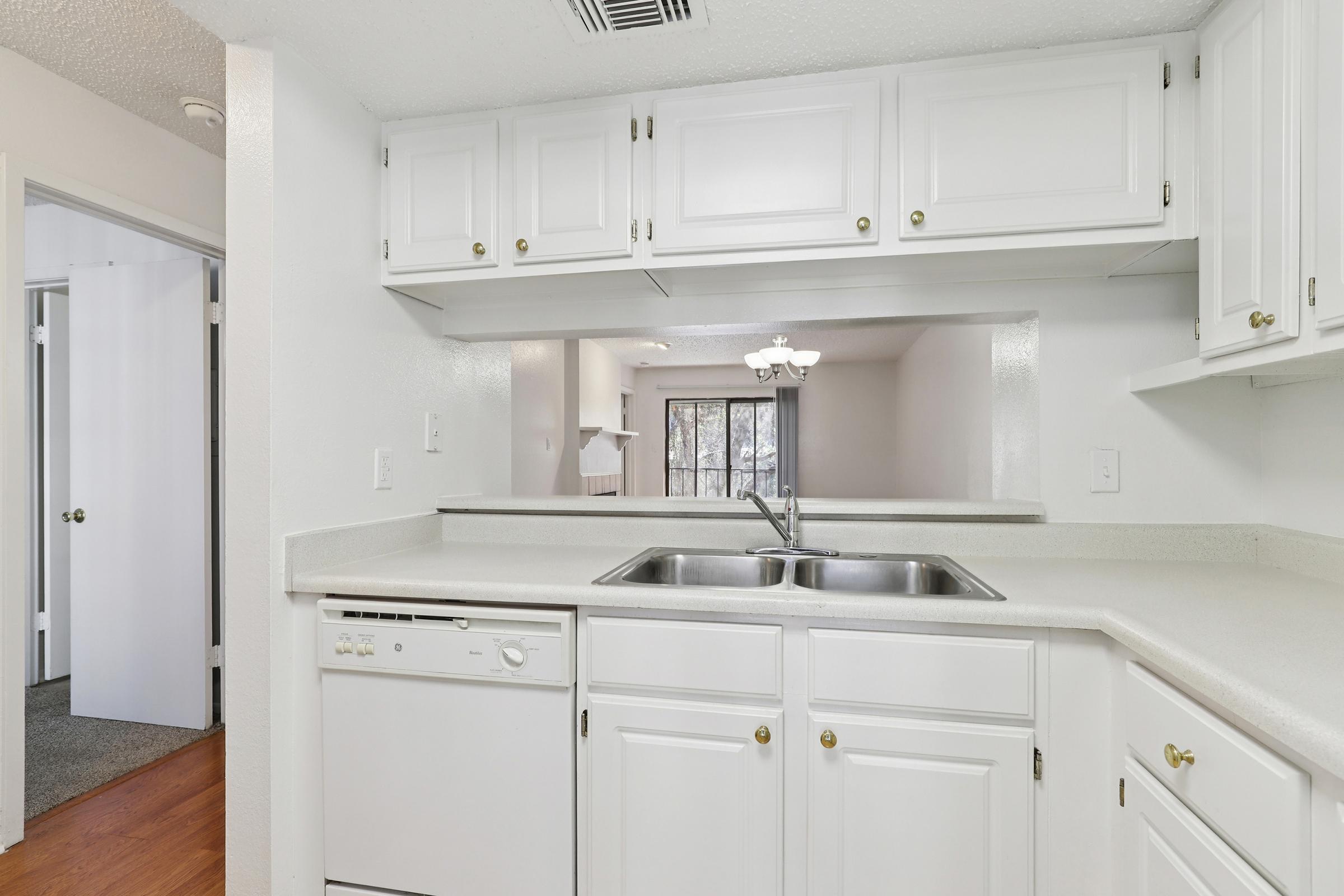 A bright, modern kitchen with white cabinets, a double sink, and a dishwasher. The countertop is light-colored, and there is an open pass-through to a living area in the background. Soft, neutral colors create a clean and inviting atmosphere, with a doorway leading to another room visible in the background.