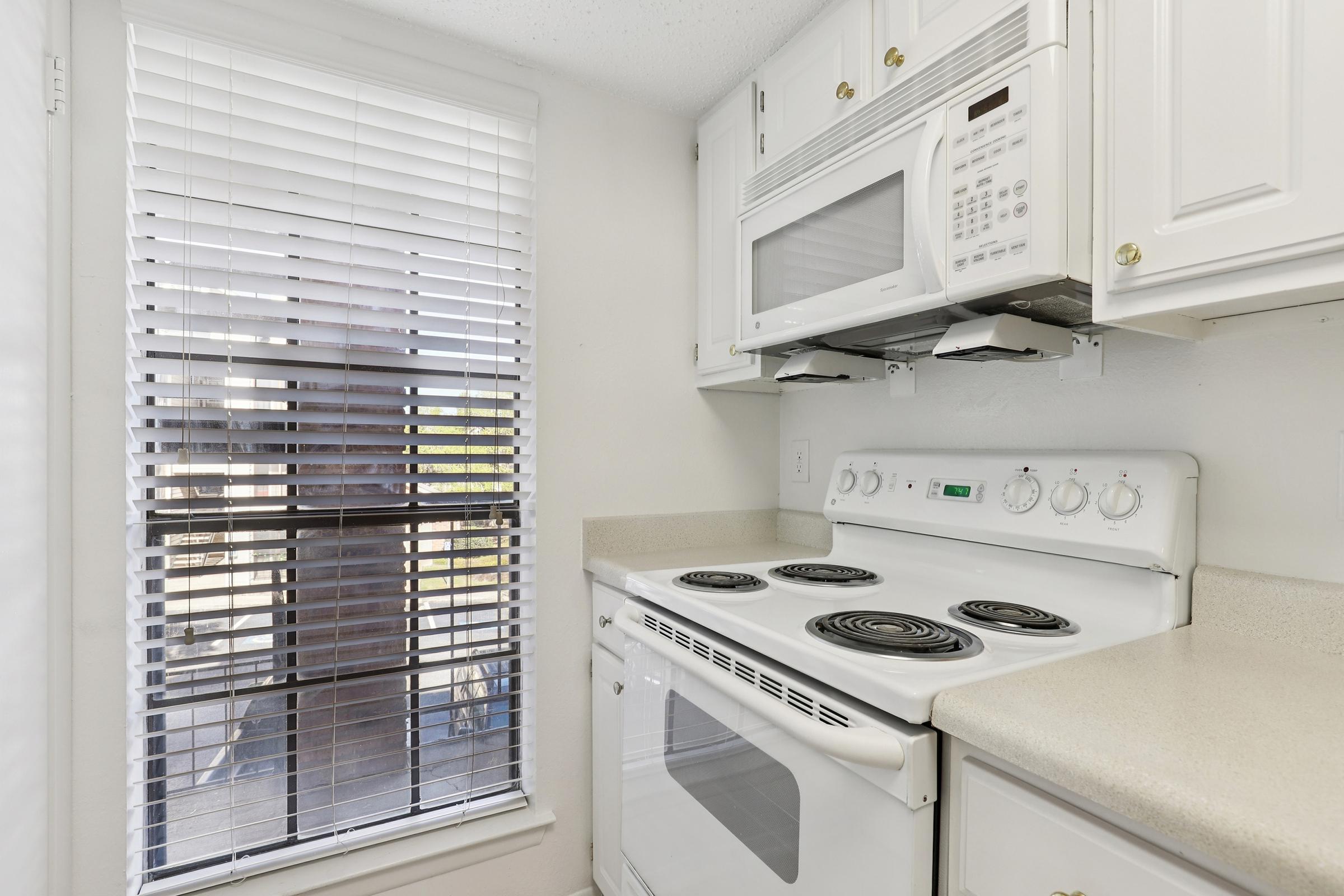 A bright kitchen featuring a white stove and microwave installed above it, with a countertop and fitted cabinetry. Natural light streams in through a window adorned with blinds, illuminating the space. The clean design highlights an efficient layout.