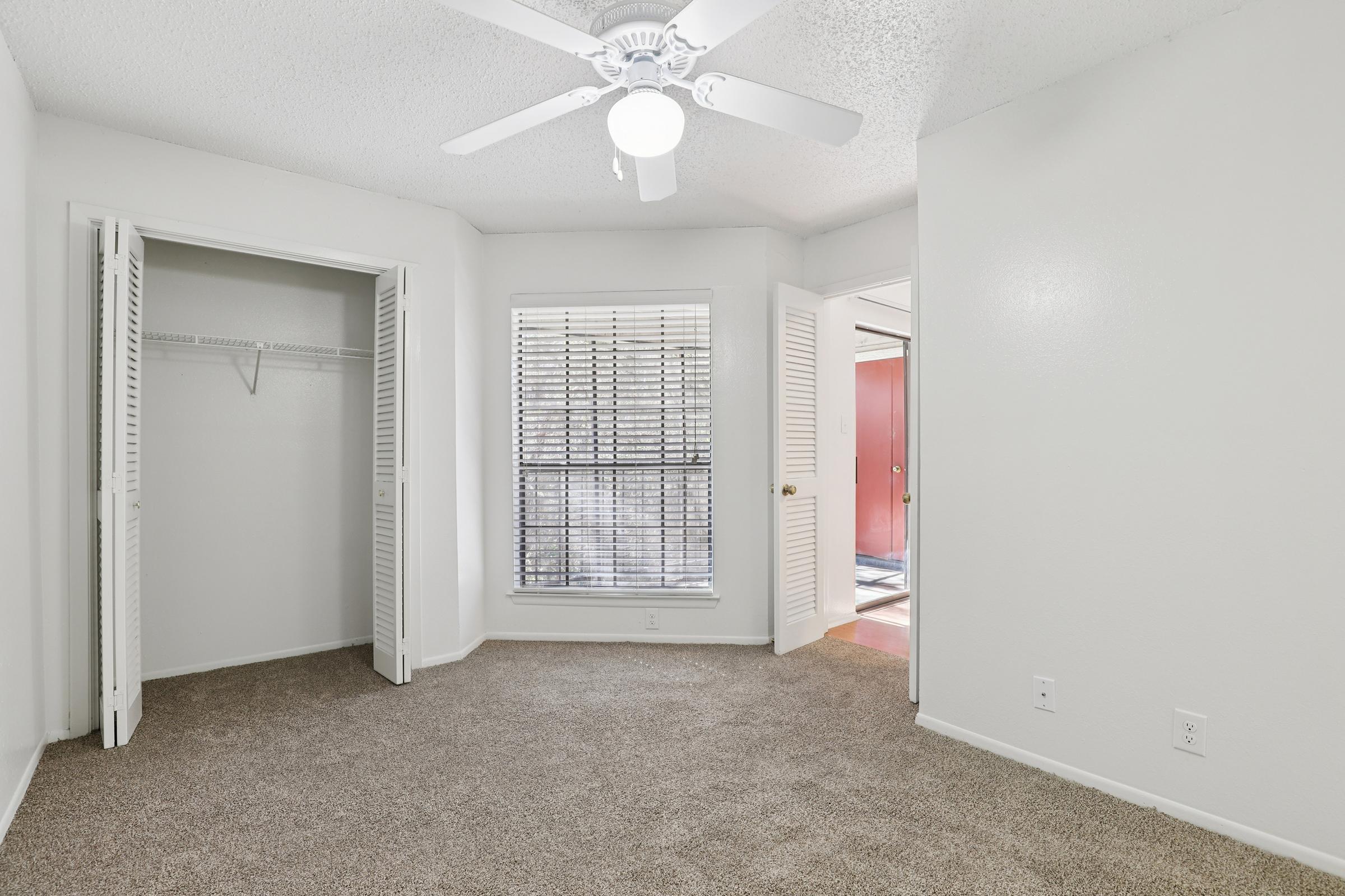 A bright, empty bedroom featuring light-colored walls and beige carpet. There is a ceiling fan, a window with blinds, and a small closet with sliding doors. The room is well-lit and has a door leading to another area.