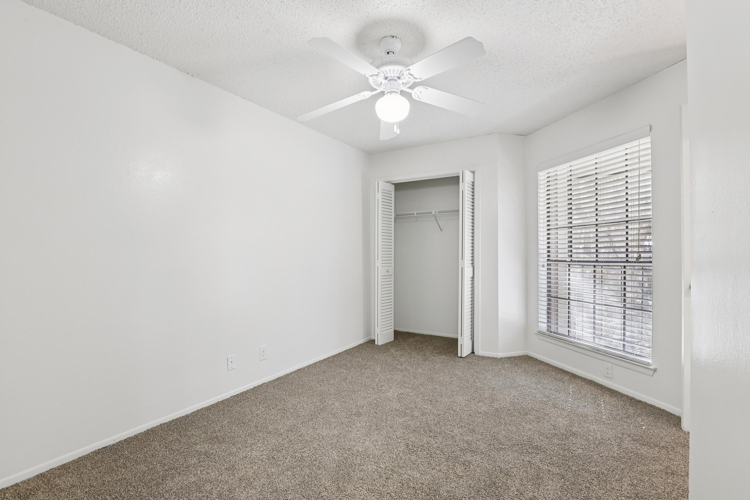 A well-lit, empty bedroom featuring light beige carpet, white walls, and a ceiling fan. It has a closet with double doors and a window with blinds, allowing natural light to enter. The room appears clean and freshly painted, making it suitable for various decor styles.