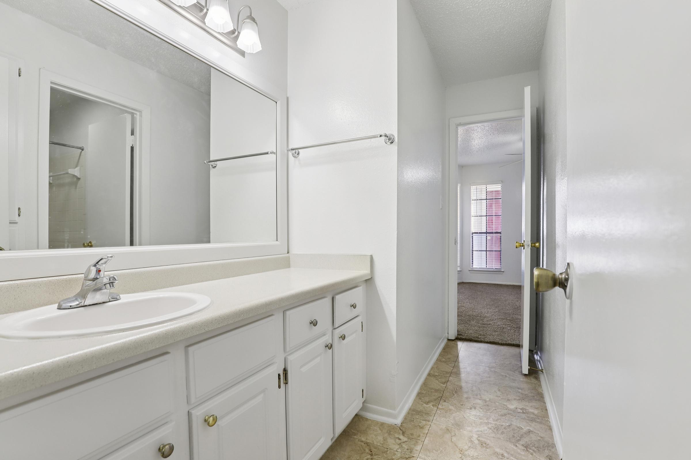 A clean and modern bathroom featuring a white countertop with a sink and mirror. The walls are painted white, and there are light fixtures above the mirror. A door in the background leads to a carpeted area with natural light coming through a window. The floor is tiled in a light color.