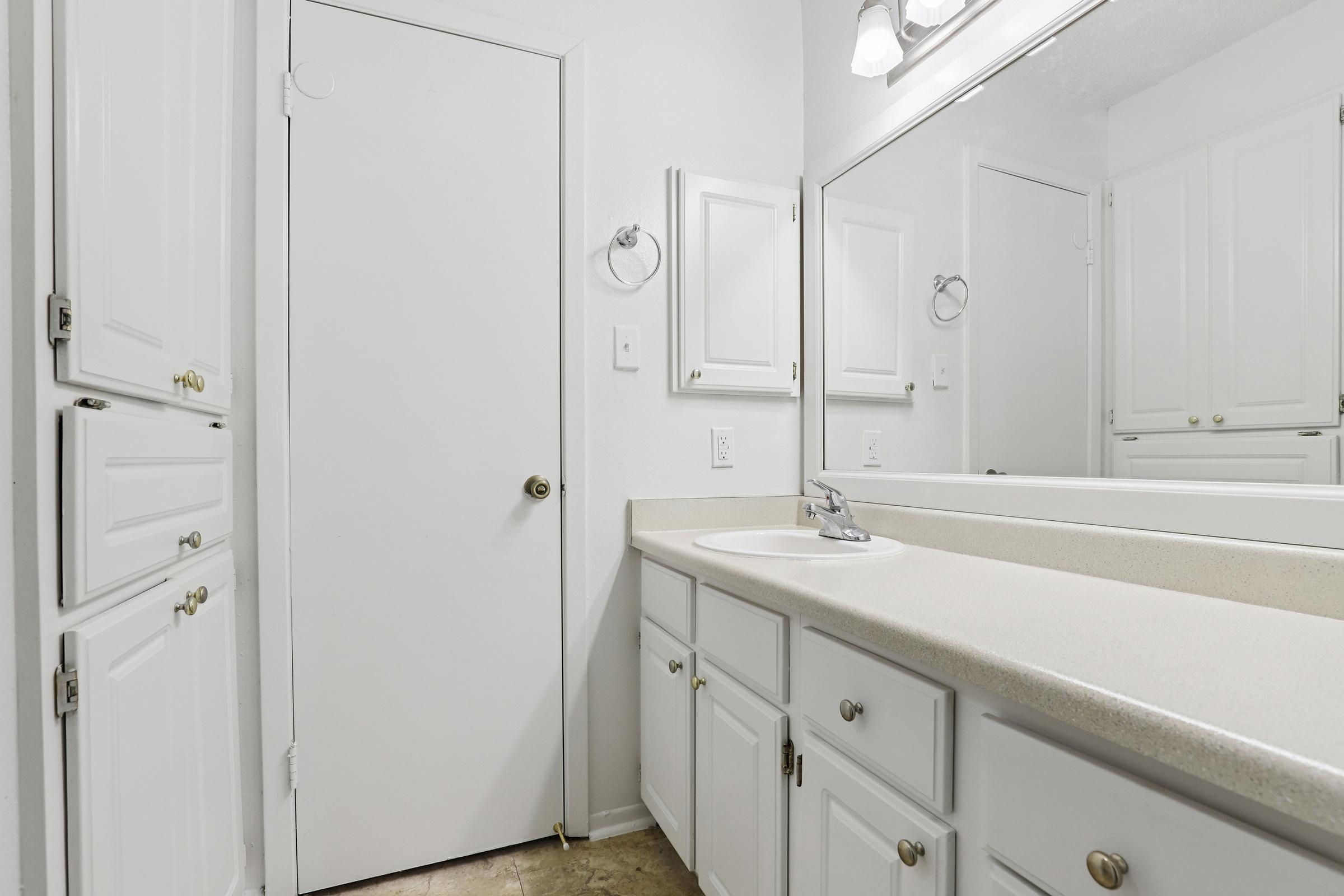 A bright, clean bathroom featuring white cabinetry, a smooth countertop, and a large mirror. A door is visible at the end of the room, and the walls are painted in a light color, enhancing the spacious feel. The overall setting is well-lit and tidy, conveying a sense of freshness.