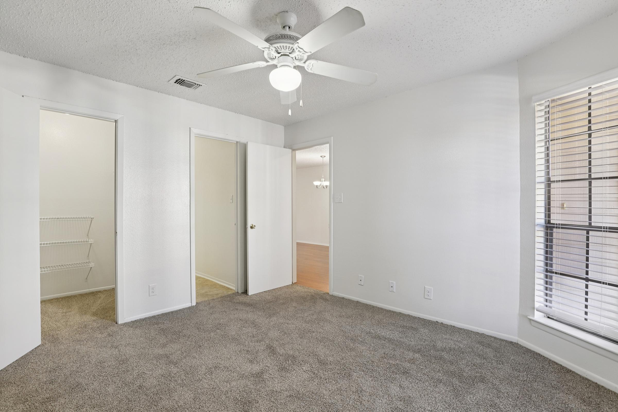 A spacious, well-lit bedroom featuring carpeted flooring and a ceiling fan. Two open doors lead to a closet and another room, while a window provides natural light, revealing white walls and simple decor. The overall ambiance is inviting and tidy.