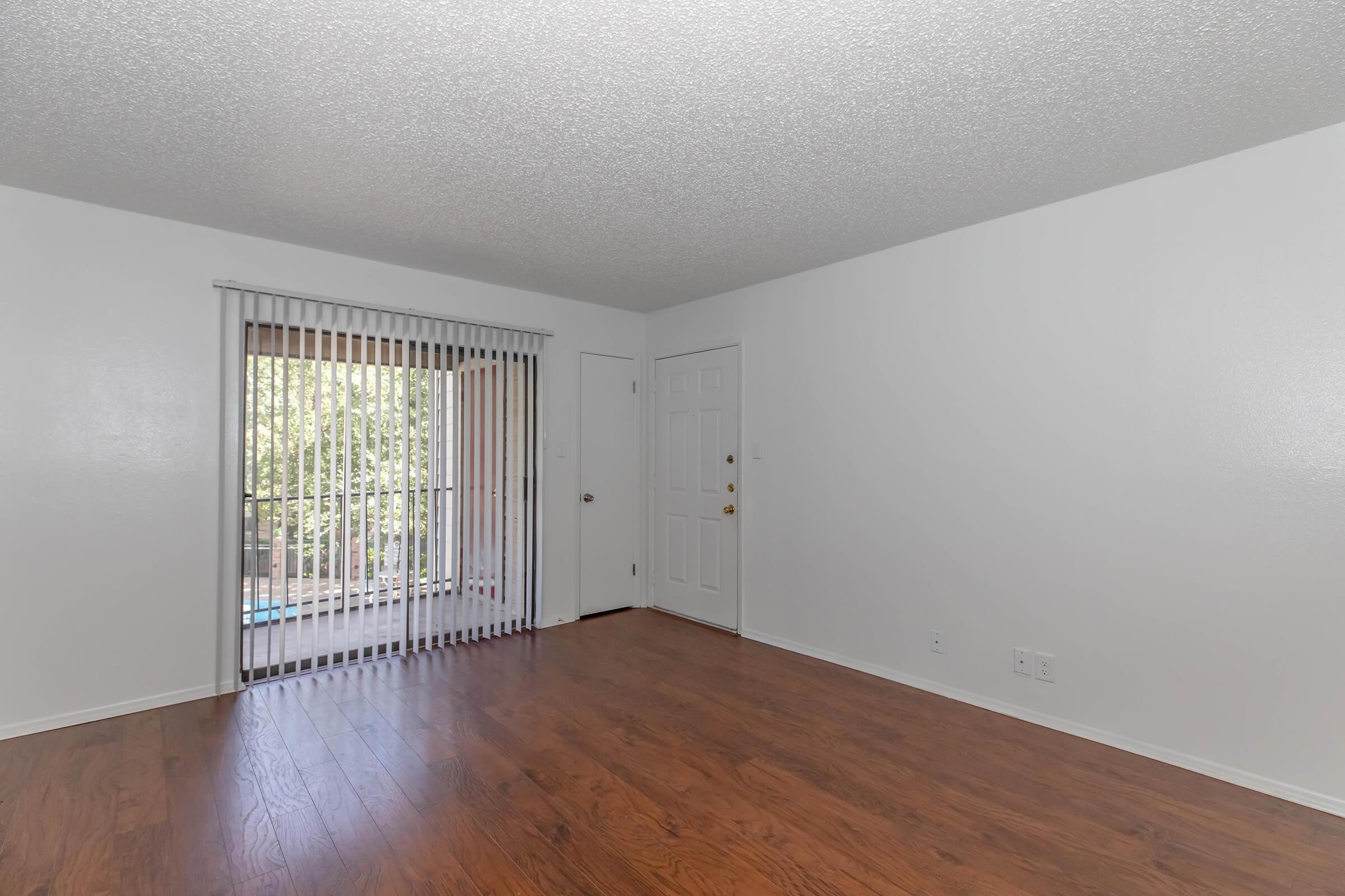 Interior view of an empty living room with wooden flooring and a sliding glass door leading to a balcony. The walls are painted white, and there are vertical blinds covering the door. A front door is visible on the right side of the image. The space is well-lit with natural light.