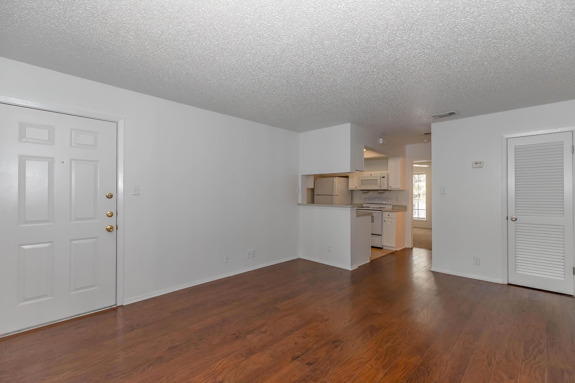 Interior view of a bright, empty room with hardwood flooring, featuring a front door on the left, an entrance to a kitchen area in the back, and a closet door on the right. The walls are painted white, and there's a textured ceiling. Natural light is coming from the kitchen area.