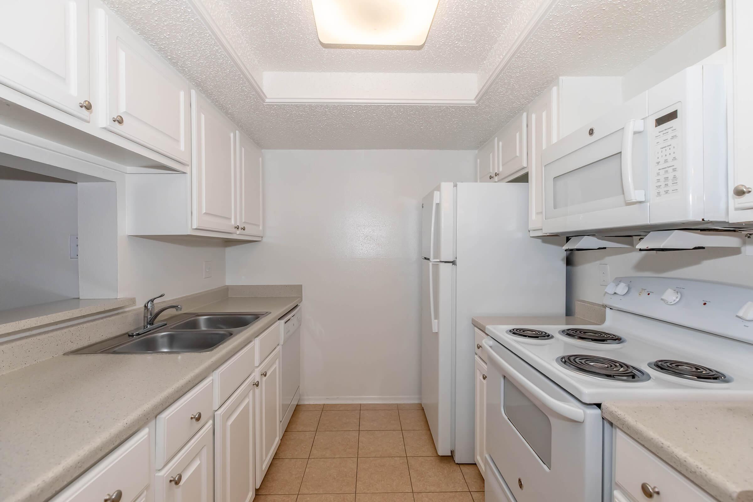 A clean, modern kitchen featuring white cabinets, a double sink, a refrigerator, a microwave, and a stove. The countertops are light-colored, and the tile floor is neutral. A small window and bright overhead lighting enhance the space.