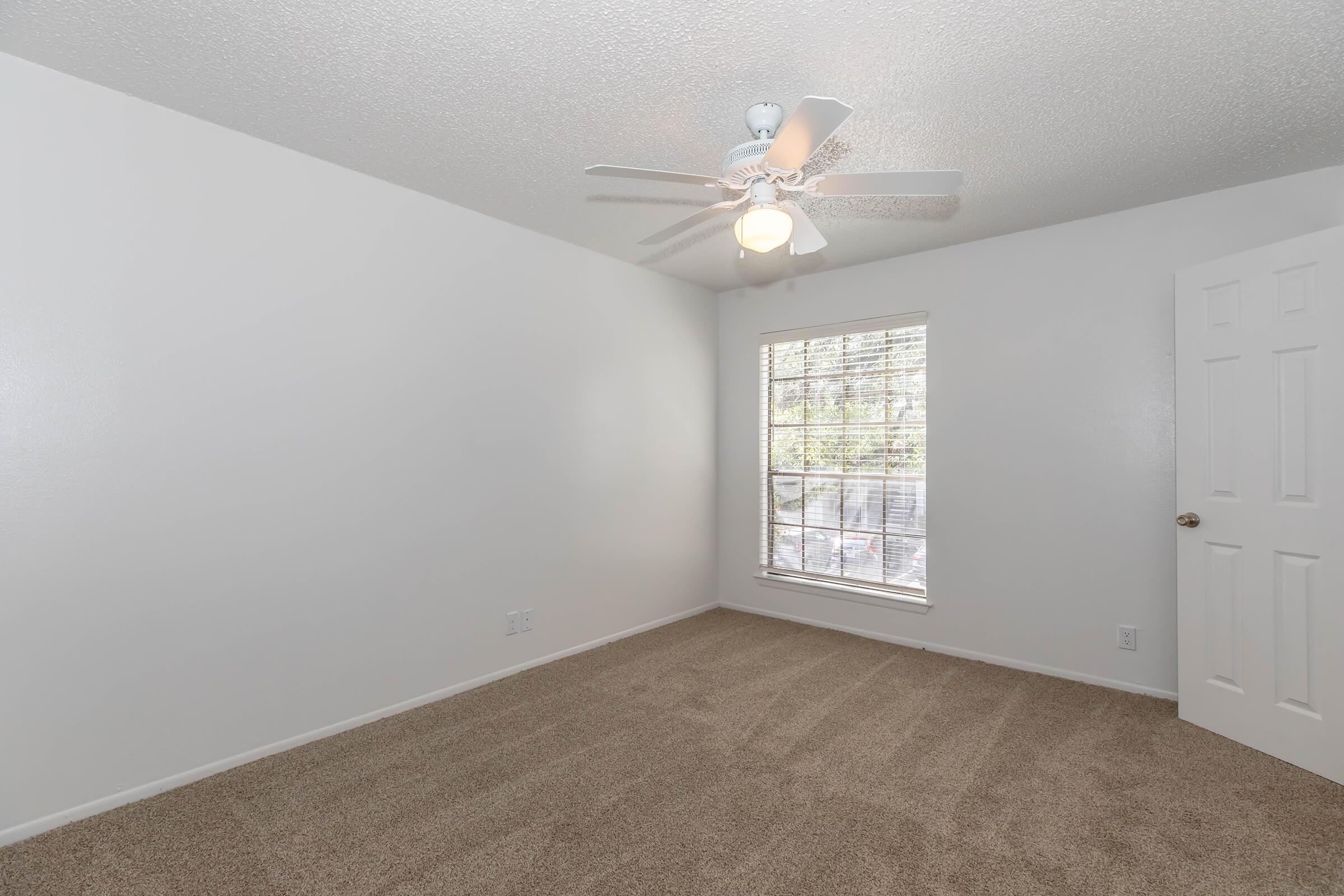 An empty bedroom featuring light-colored walls, a window with blinds allowing natural light, a ceiling fan, and carpeted flooring. The door is slightly ajar, and the space appears clean and well-lit, making it suitable for various decorating styles.