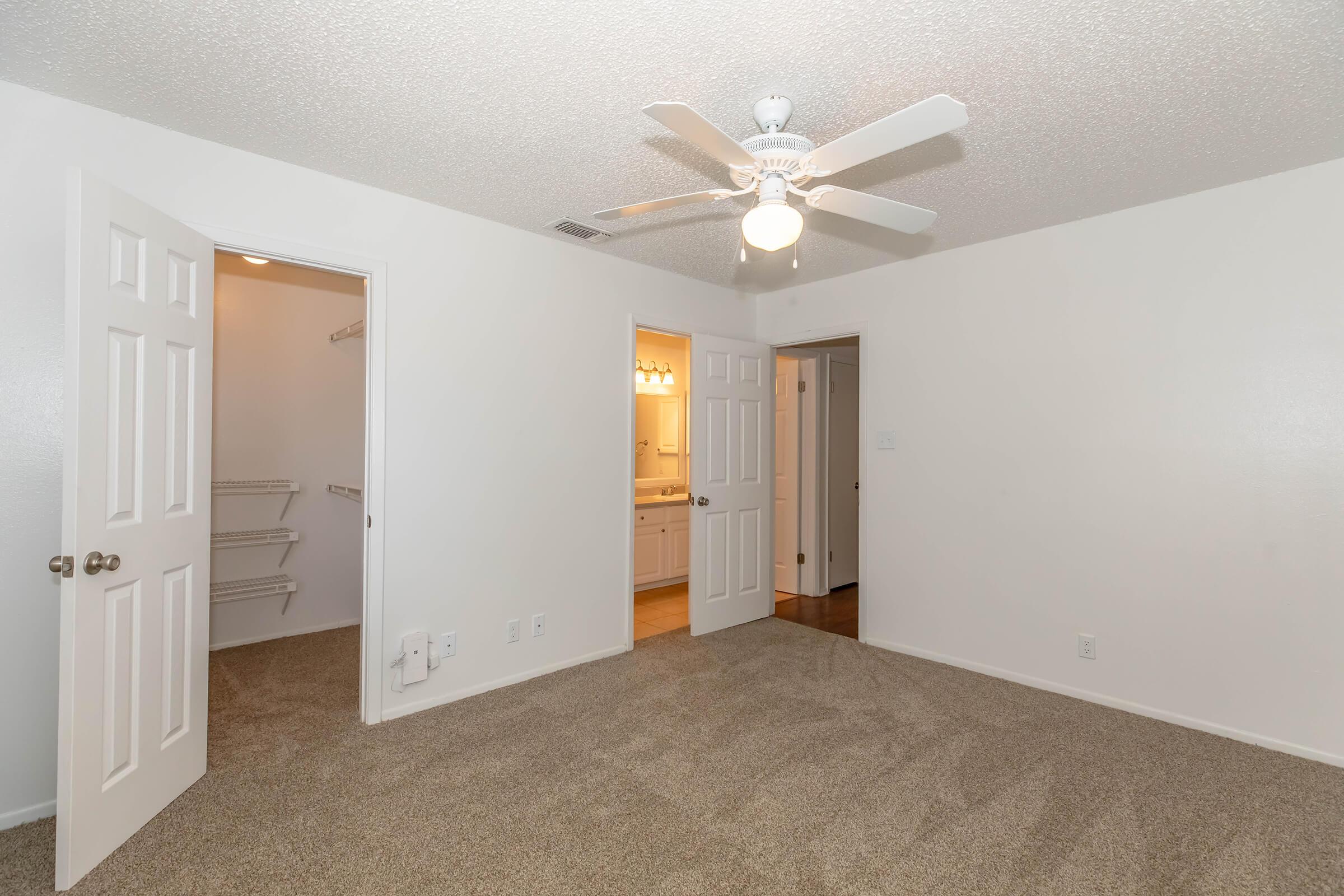 A bright, empty bedroom features light-colored walls and a ceiling fan. Two doors lead to a closet and a bathroom. The floor is covered in beige carpeting, creating a cozy atmosphere. Natural light appears to be coming from the bathroom area.