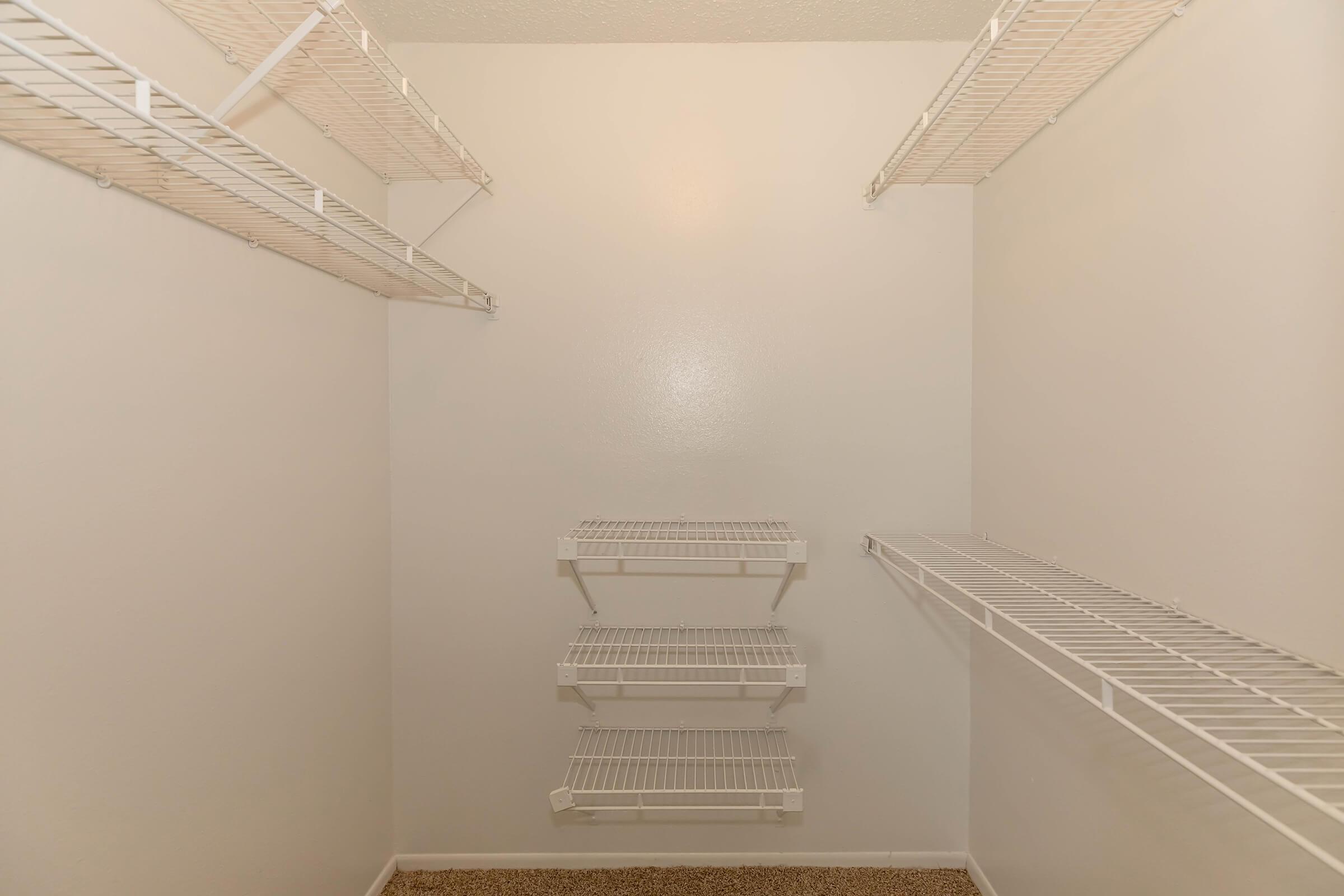Empty walk-in closet featuring white wire shelving on all sides, with three shelves in the center and additional shelves on the left and right. The walls are painted white, and the flooring is carpeted in a neutral color. The overall space is bright and spacious, with minimal decor.