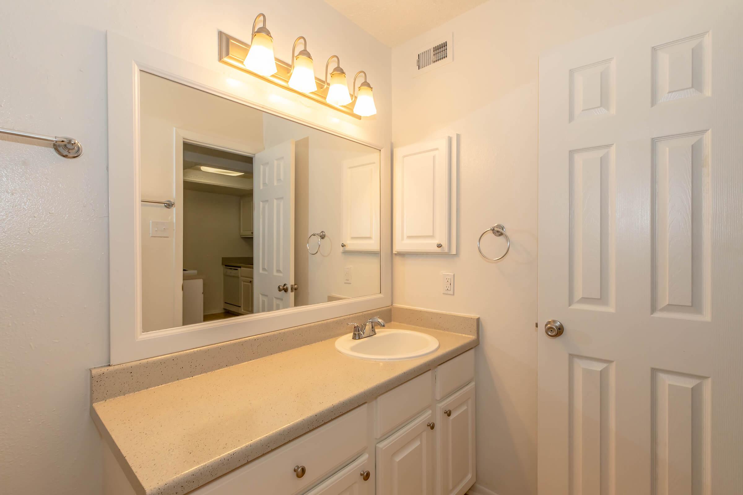 Bright bathroom featuring a double vanity with a light-colored countertop, white cabinets, and a large mirror above. The area includes modern lighting and a towel ring on the wall. A door leads to another room, and the overall design is clean and inviting.