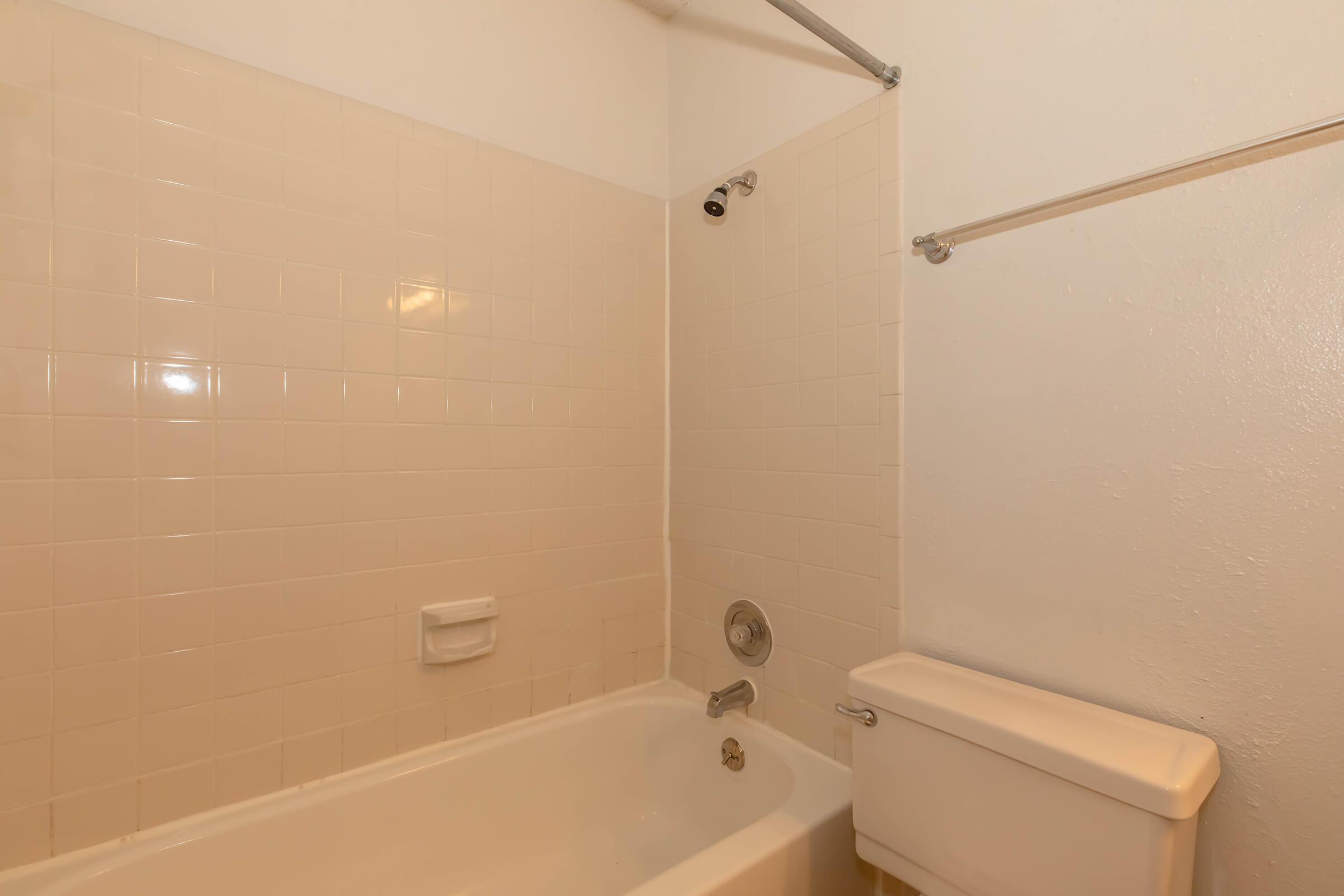 A simple bathroom with a white tiled shower and bathtub. The bathtub is on the left side, equipped with a showerhead above. A toilet is positioned next to the bathtub. The walls are painted white, and there are metal fixtures for a shower curtain. The overall appearance is clean and functional.