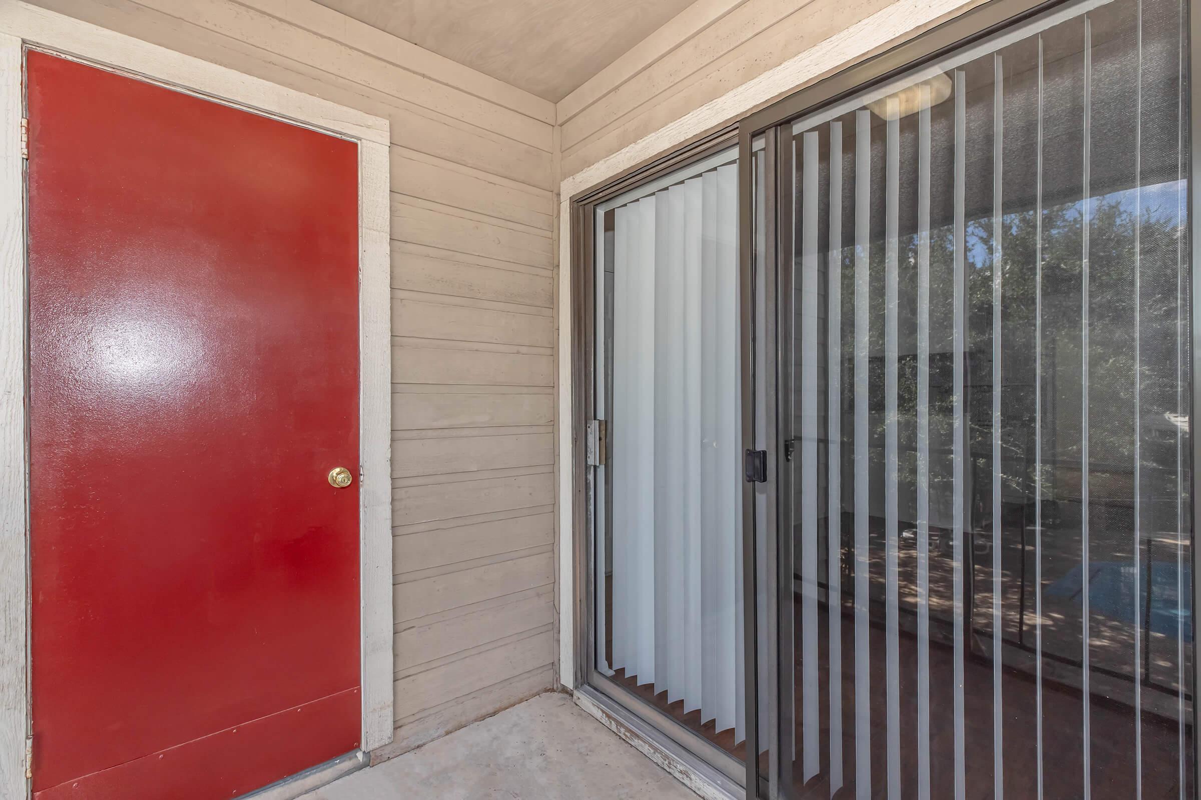 A red door on the left side of a porch entrance, with a sliding glass door on the right featuring vertical blinds. The entrance has a light-colored wall and a concrete floor, giving an inviting look to the entryway. Natural light filters in from the outside.