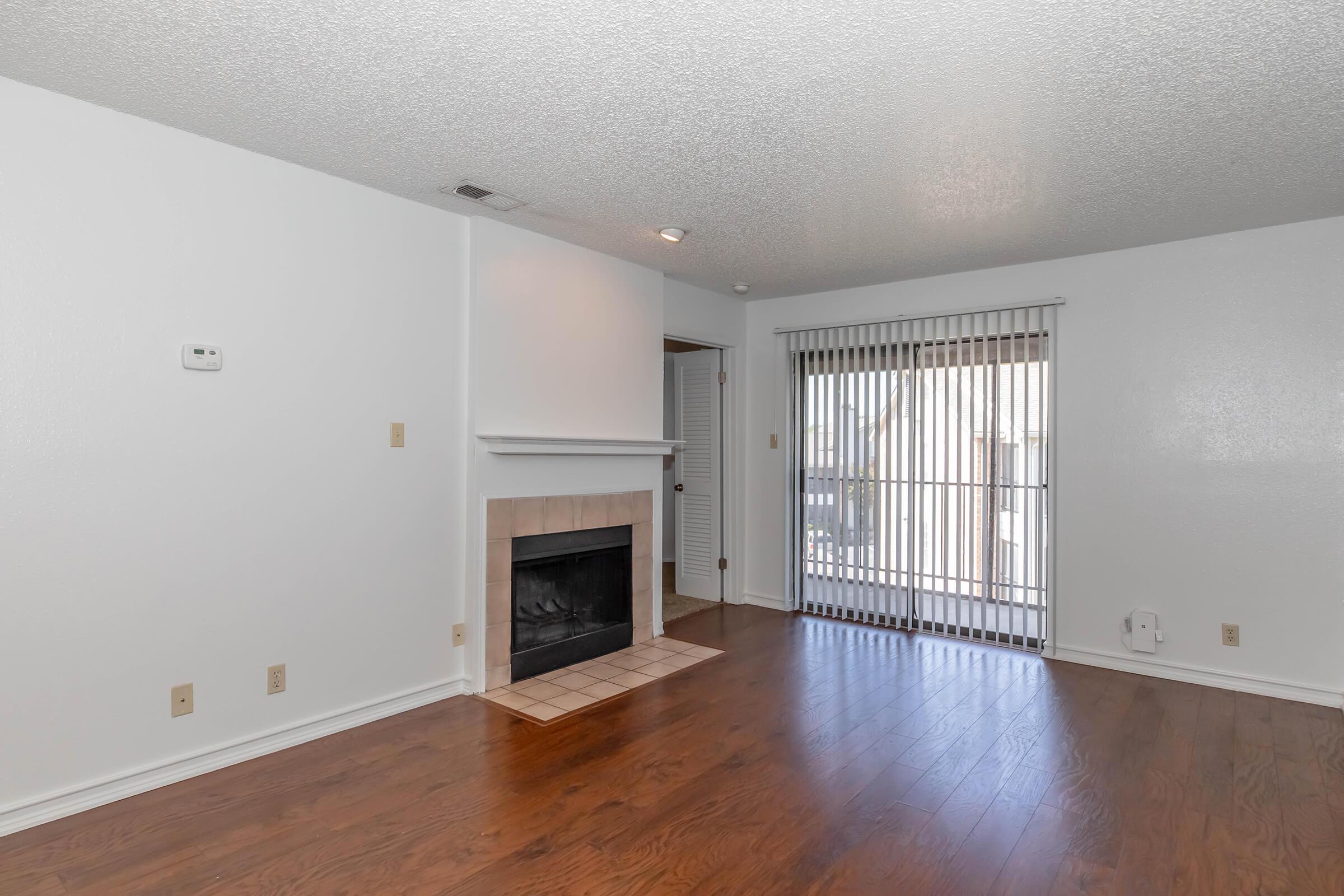 A spacious living room featuring a white textured ceiling, hardwood flooring, and a cozy fireplace with a tiled surround. Large sliding glass doors lead to an outdoor area, complemented by vertical blinds. The walls are painted white, creating a bright and airy atmosphere.
