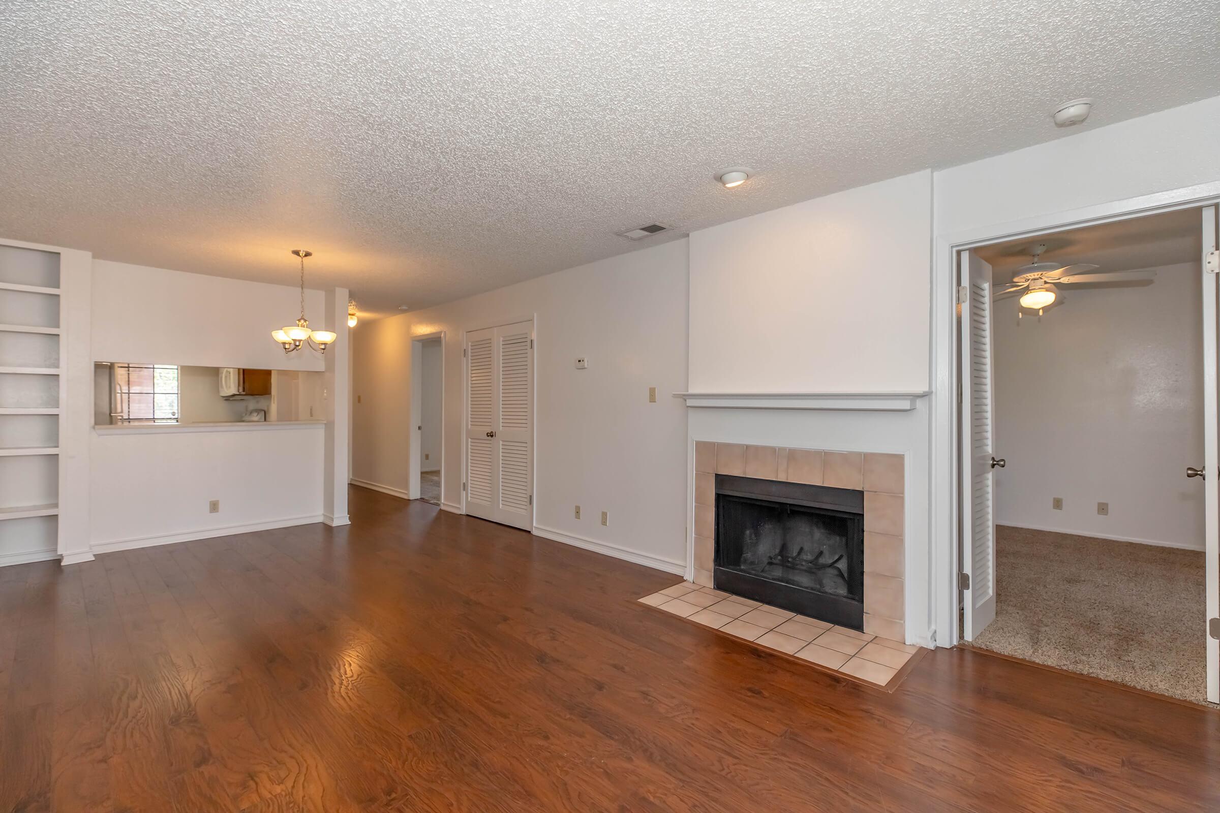Interior view of a living space featuring hardwood floors, a fireplace with a tiled surround, and a breakfast bar leading to a kitchen. The room has a light, open feel with a ceiling fan and multiple entry points, including a doorway to a carpeted area.