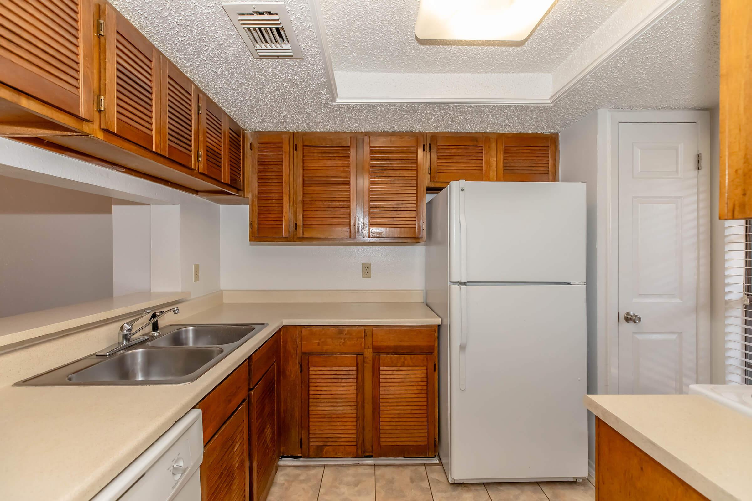 Bright kitchen featuring wooden cabinets with louvered doors, white countertops, and a double sink. A white refrigerator stands against a wall, and there are beige floor tiles. The kitchen has a clean and modern look with a simple design.