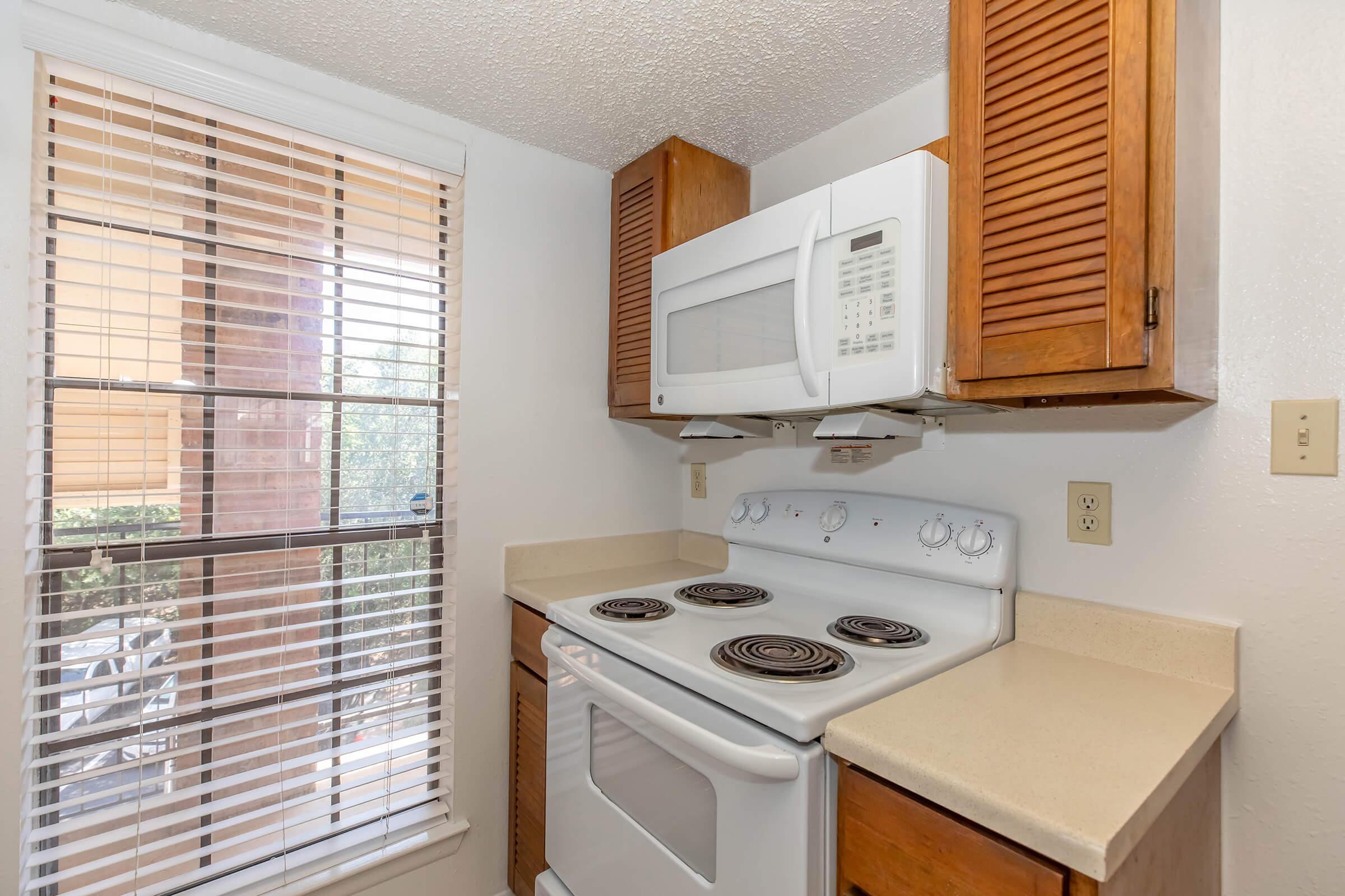 A small kitchen featuring a white stove and oven, a microwave mounted above, and wooden cabinets. There is a window with blinds allowing natural light to enter, and a light-colored countertop underneath the appliances. The setting is clean and organized, typical of an apartment kitchen.