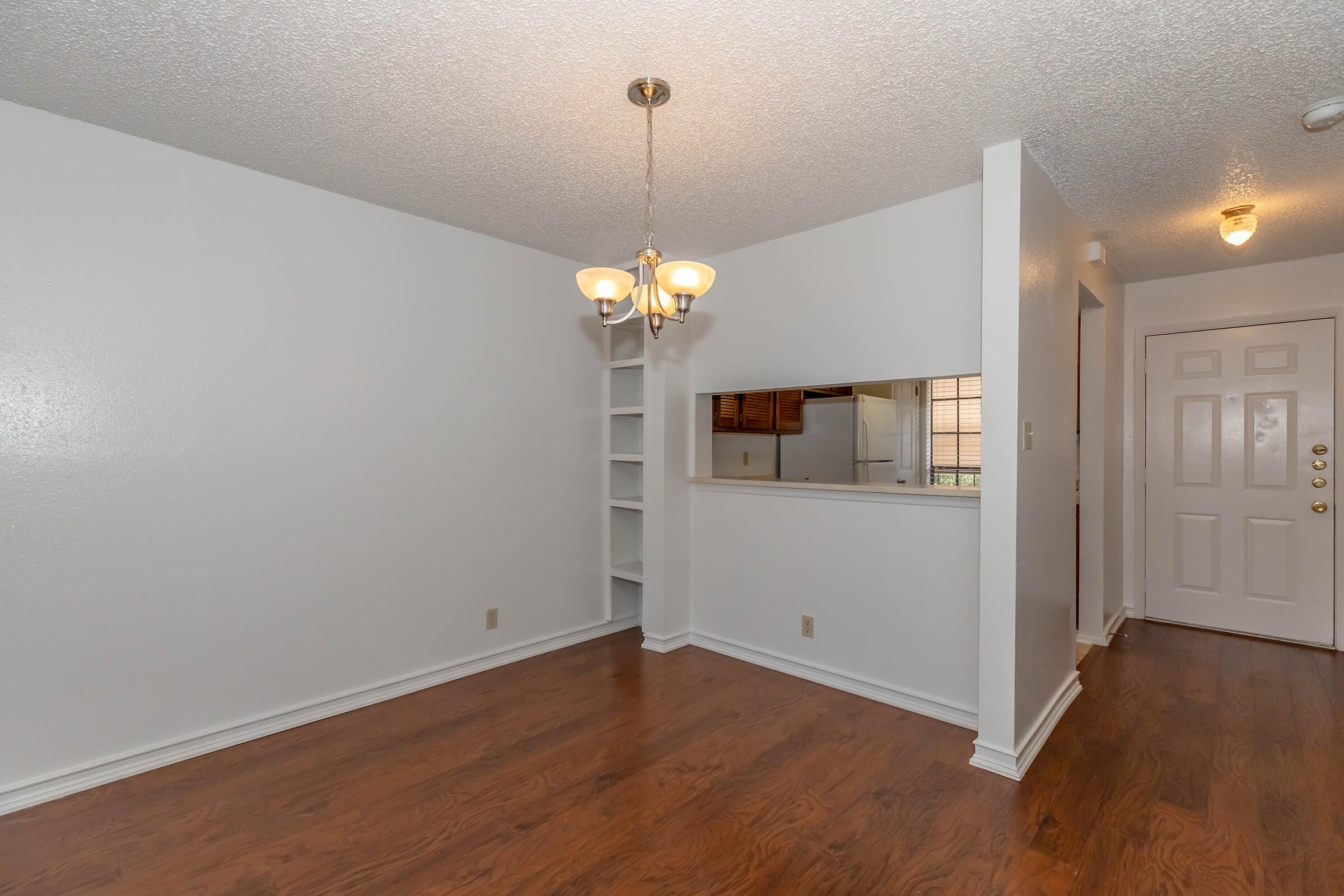 A well-lit dining area featuring a chandelier, light-colored walls, and hardwood flooring. To the left, there are built-in shelves, and a pass-through window leading into the kitchen area. A front door is visible on the right side of the image. The space has a clean and open layout.