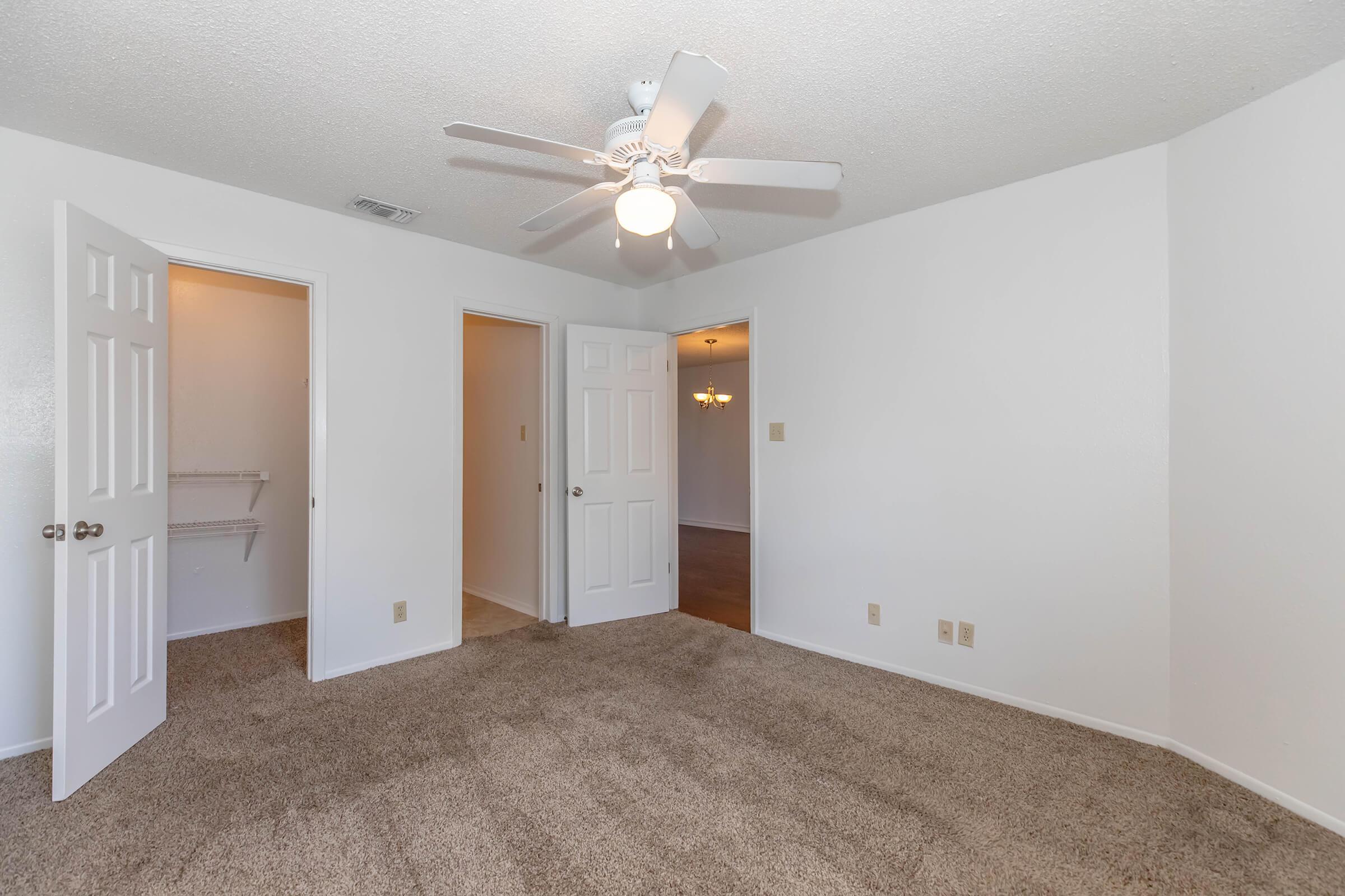 A well-lit, empty bedroom featuring beige carpet, white walls, and a ceiling fan. There are two doors leading to closets and another door opening to an adjoining room. Natural light fills the space, emphasizing the neutral color palette and clean, airy feel of the room.