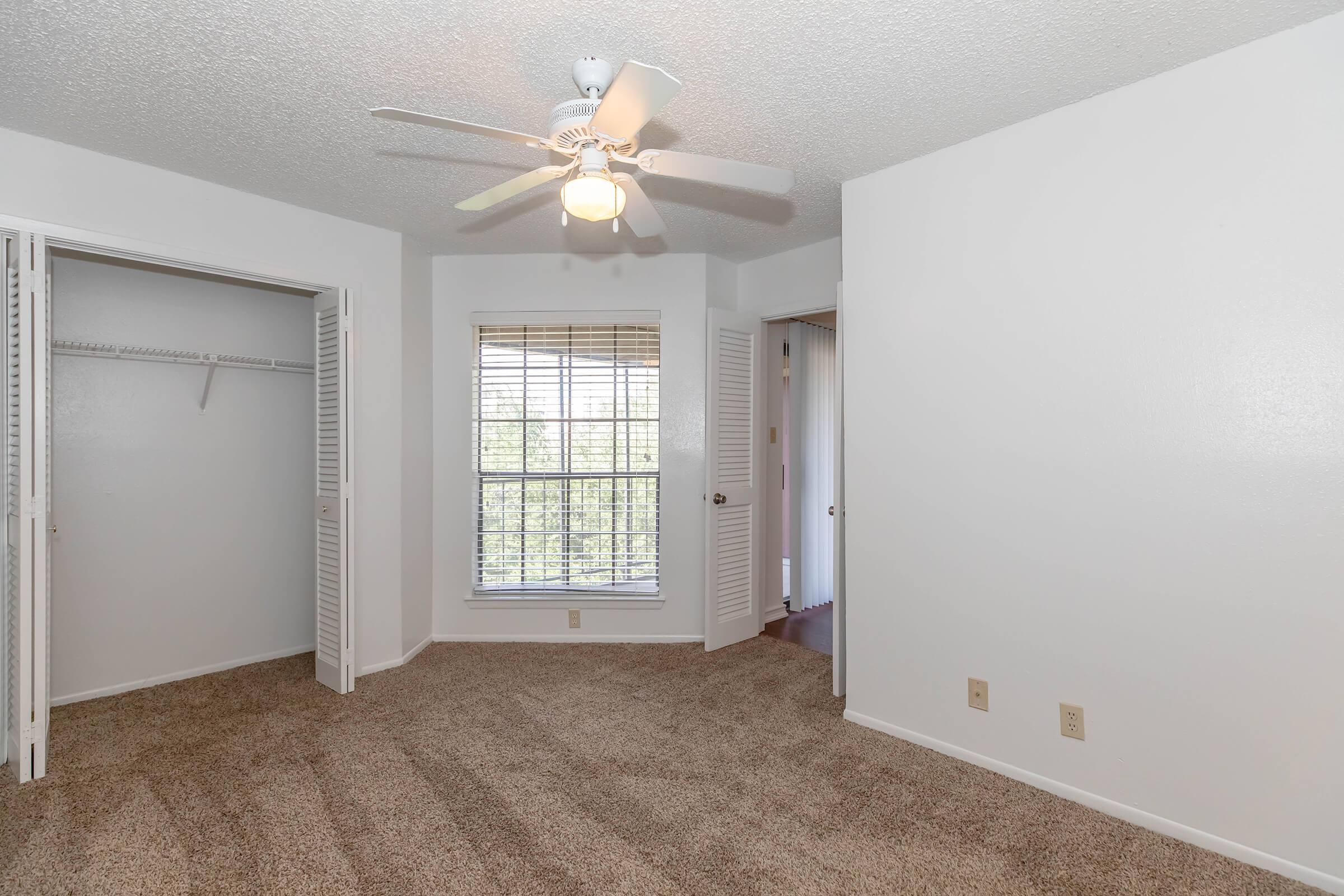 A spacious bedroom featuring beige carpet, a ceiling fan, and large windows with blinds. The room has a closet with sliding doors on one side and a doorway leading to another area. The walls are painted white, creating a bright and airy atmosphere.