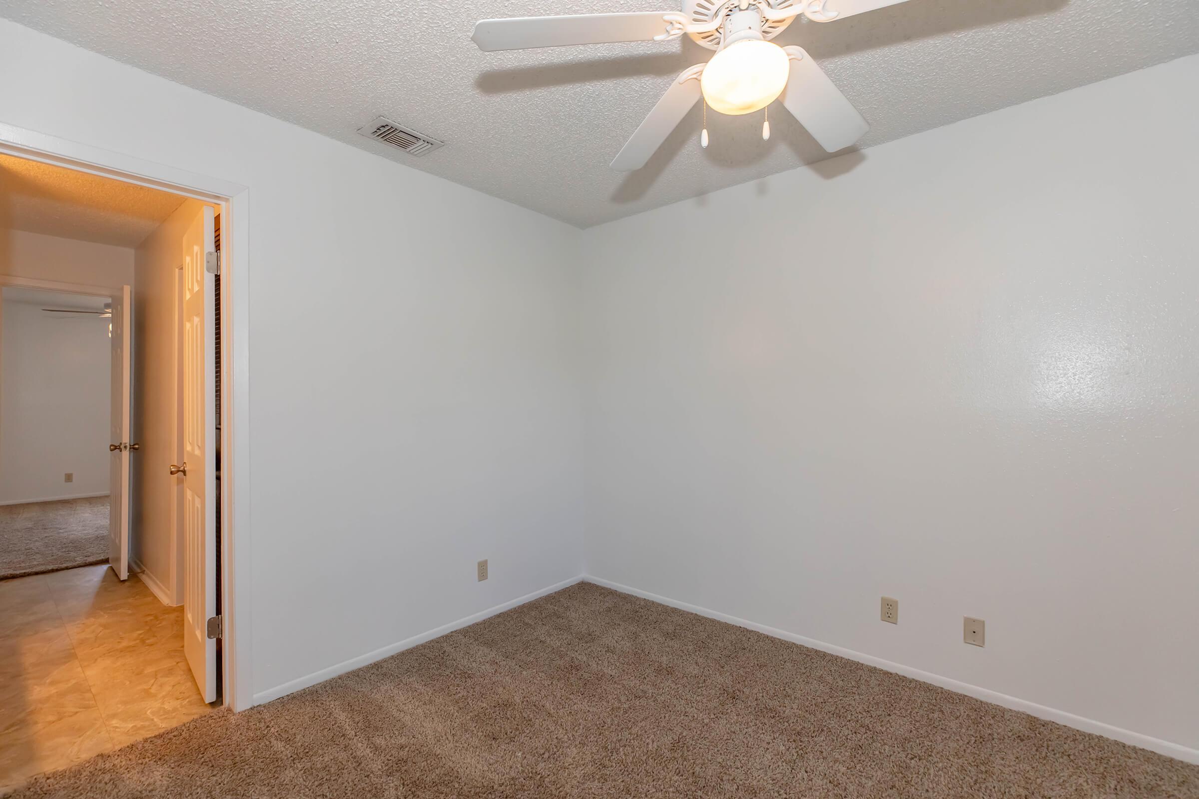 A vacant room featuring beige carpet flooring, white walls, and a ceiling fan. There is a doorway leading to another room in the background. The space has no furniture and is well-lit, creating a bright and open atmosphere.
